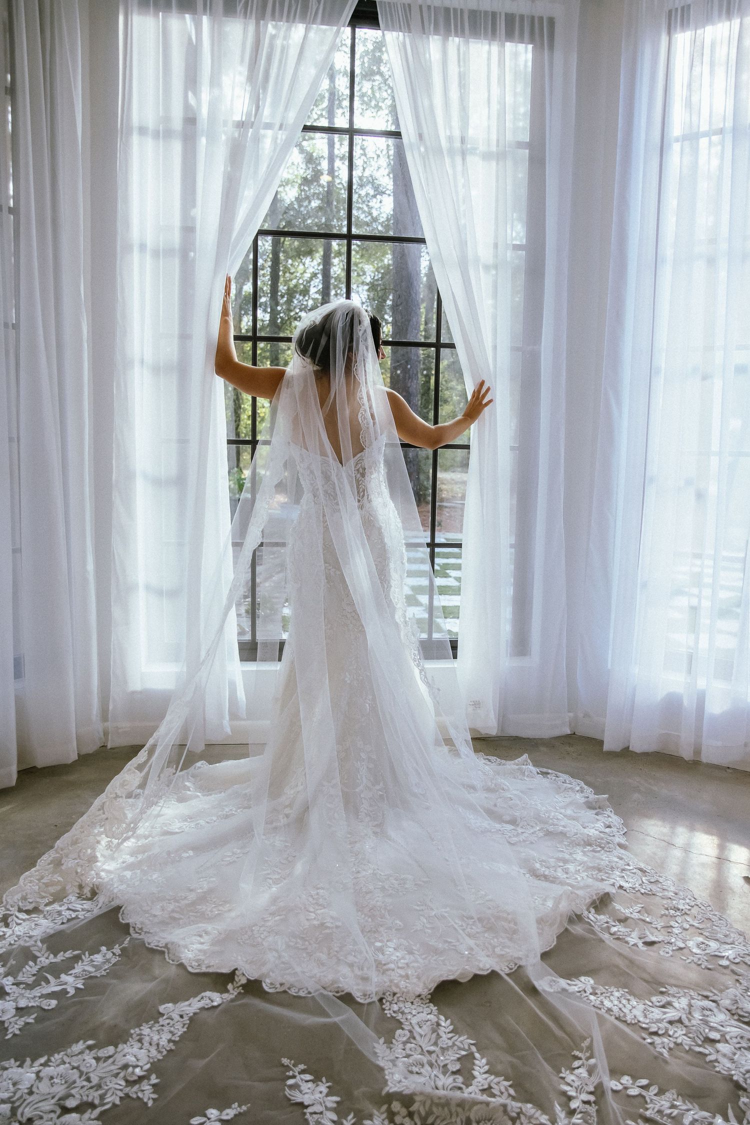 Bride in lace wedding gown and cathedral veil standing before a large window with white curtains.