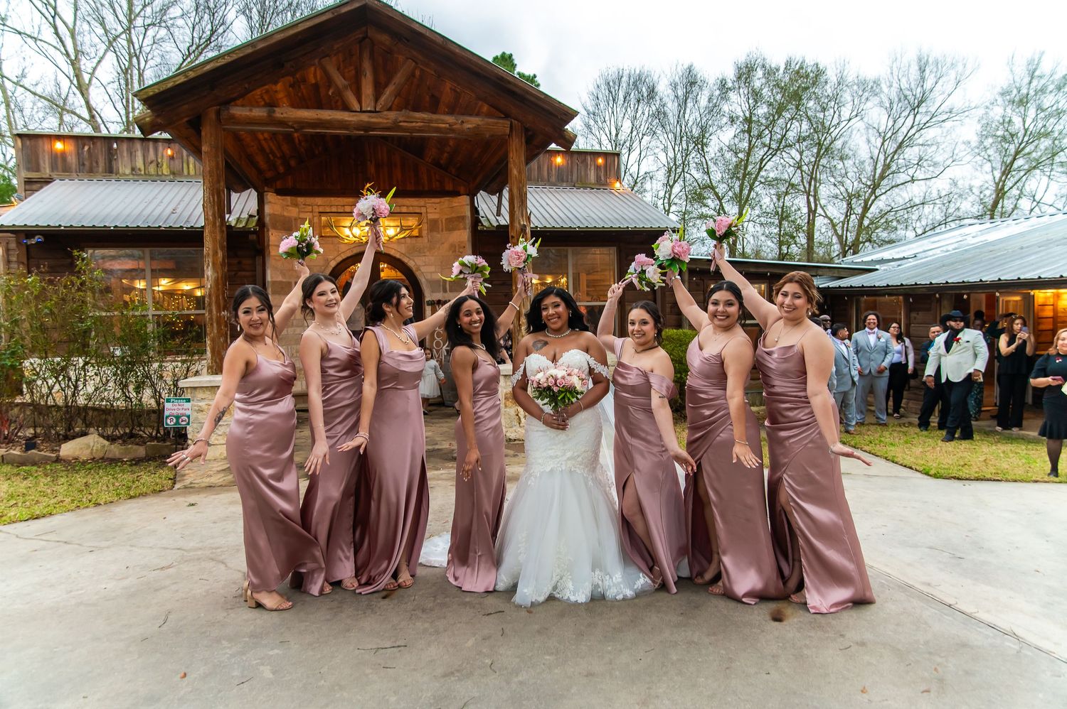 Bride in white gown poses with eight bridesmaids in dusty rose dresses, all cheering outside rustic venue.