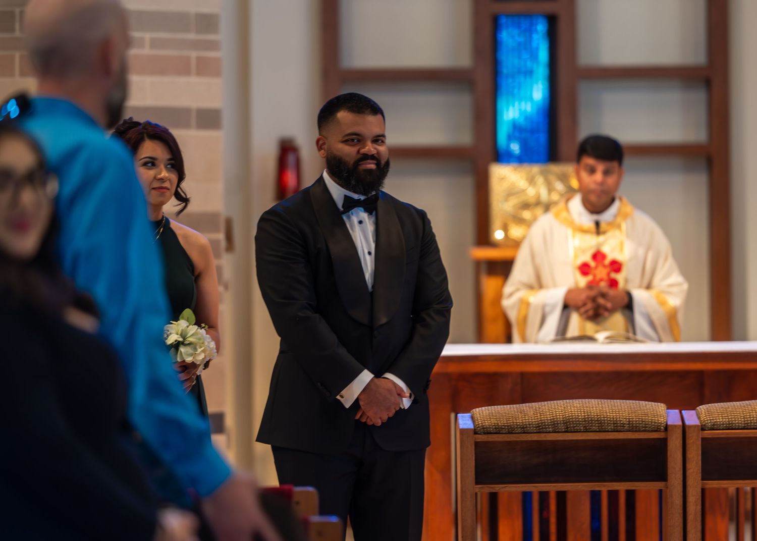 Groom in black tuxedo smiling as bride walks down church aisle during wedding ceremony.
