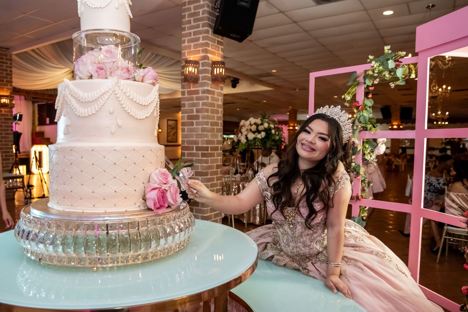 Quinceañera girl in pink gown cutting a tall elegant white wedding-style cake at her celebration.