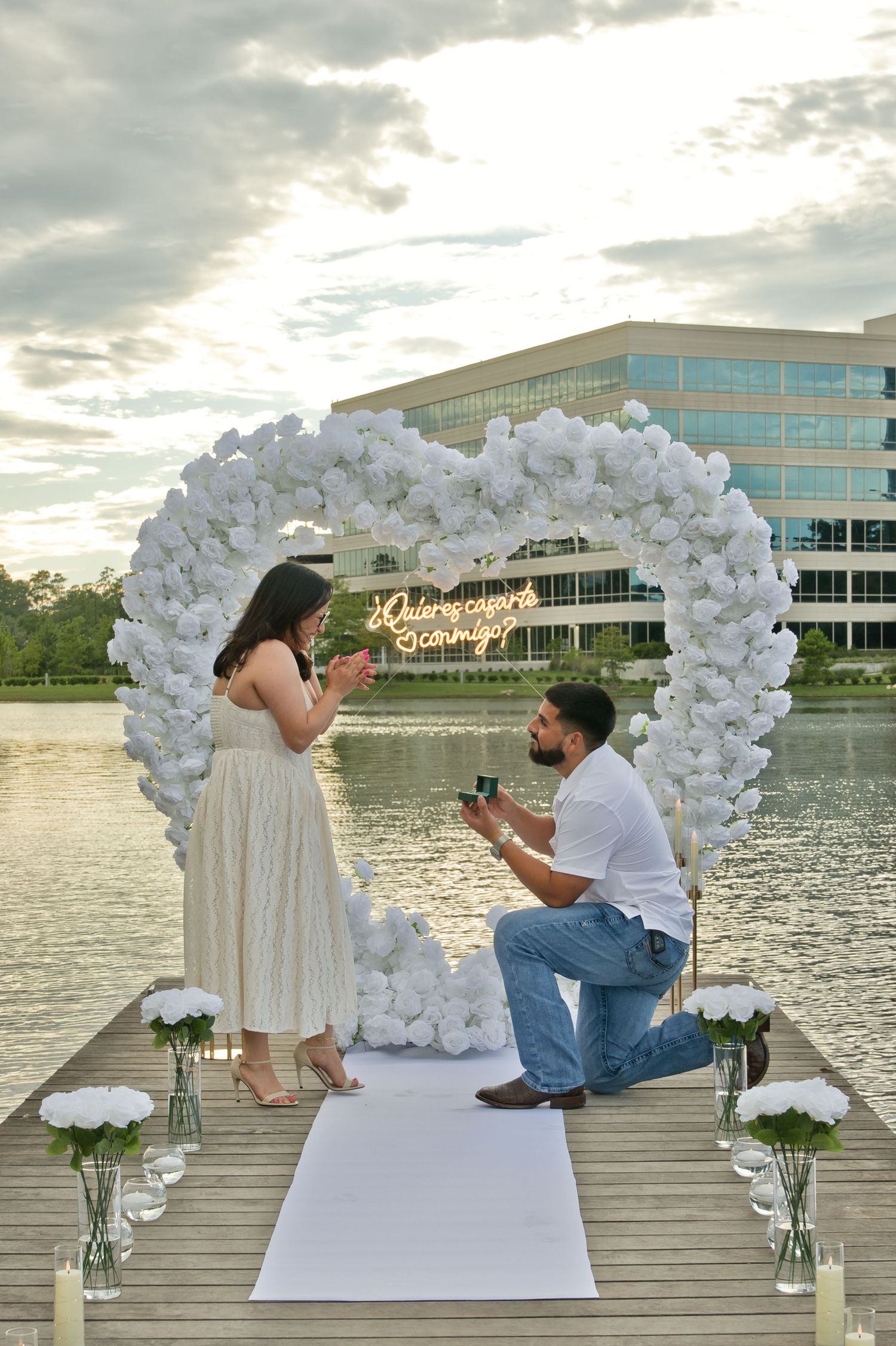 Man proposing on knee under white floral heart arch on lake dock at dusk.