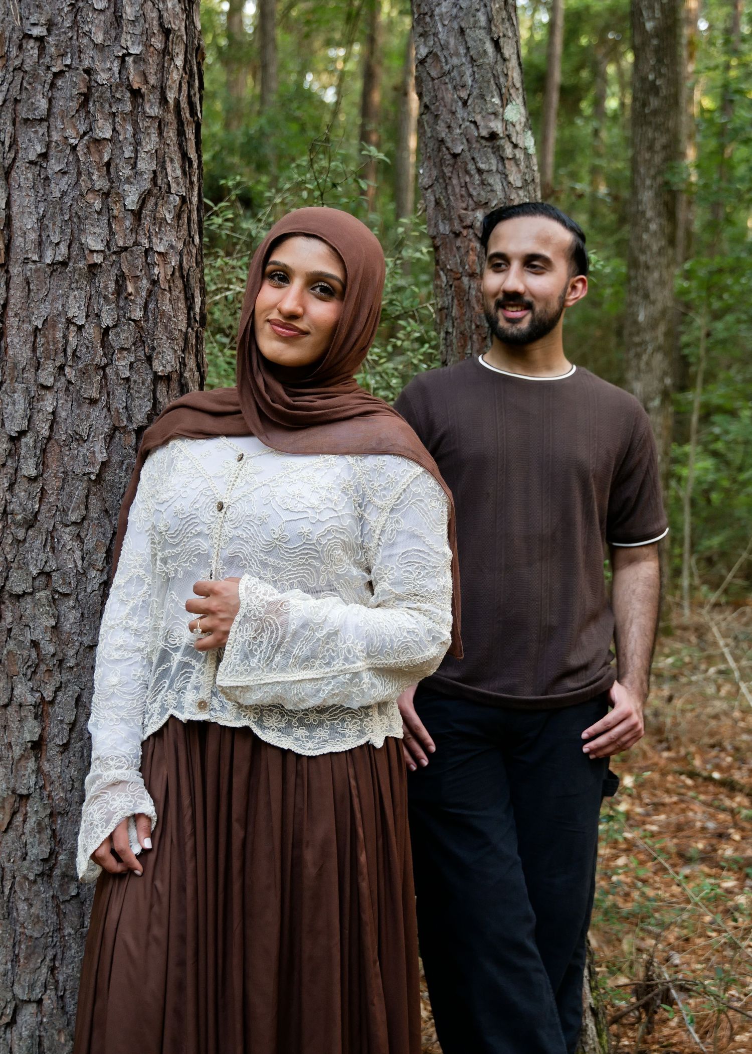 Couple posing in forest, woman in hijab and lace top, man in brown shirt.
