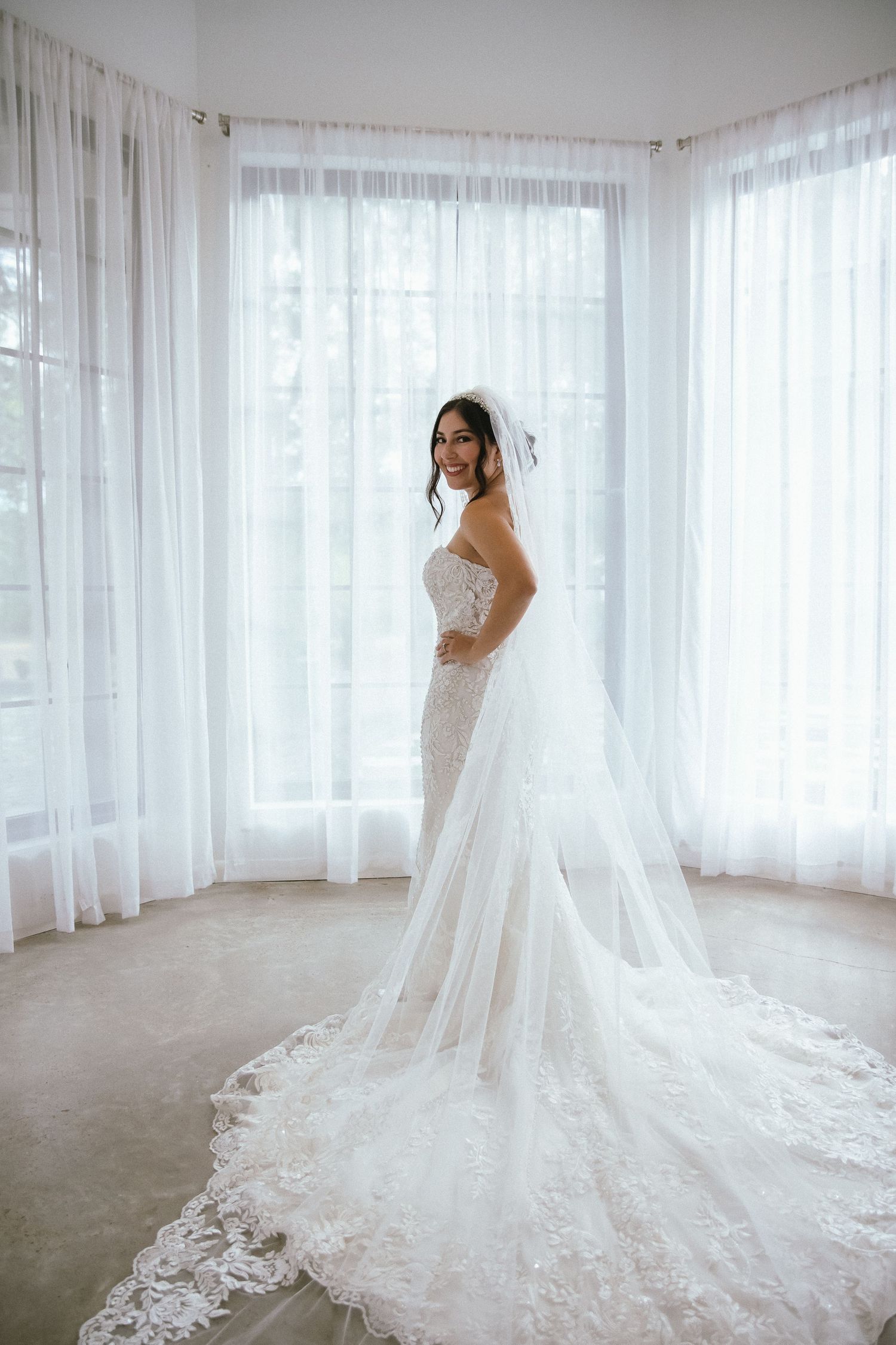 Bride in strapless lace mermaid wedding gown with long cathedral train and veil stands before sheer white curtains.