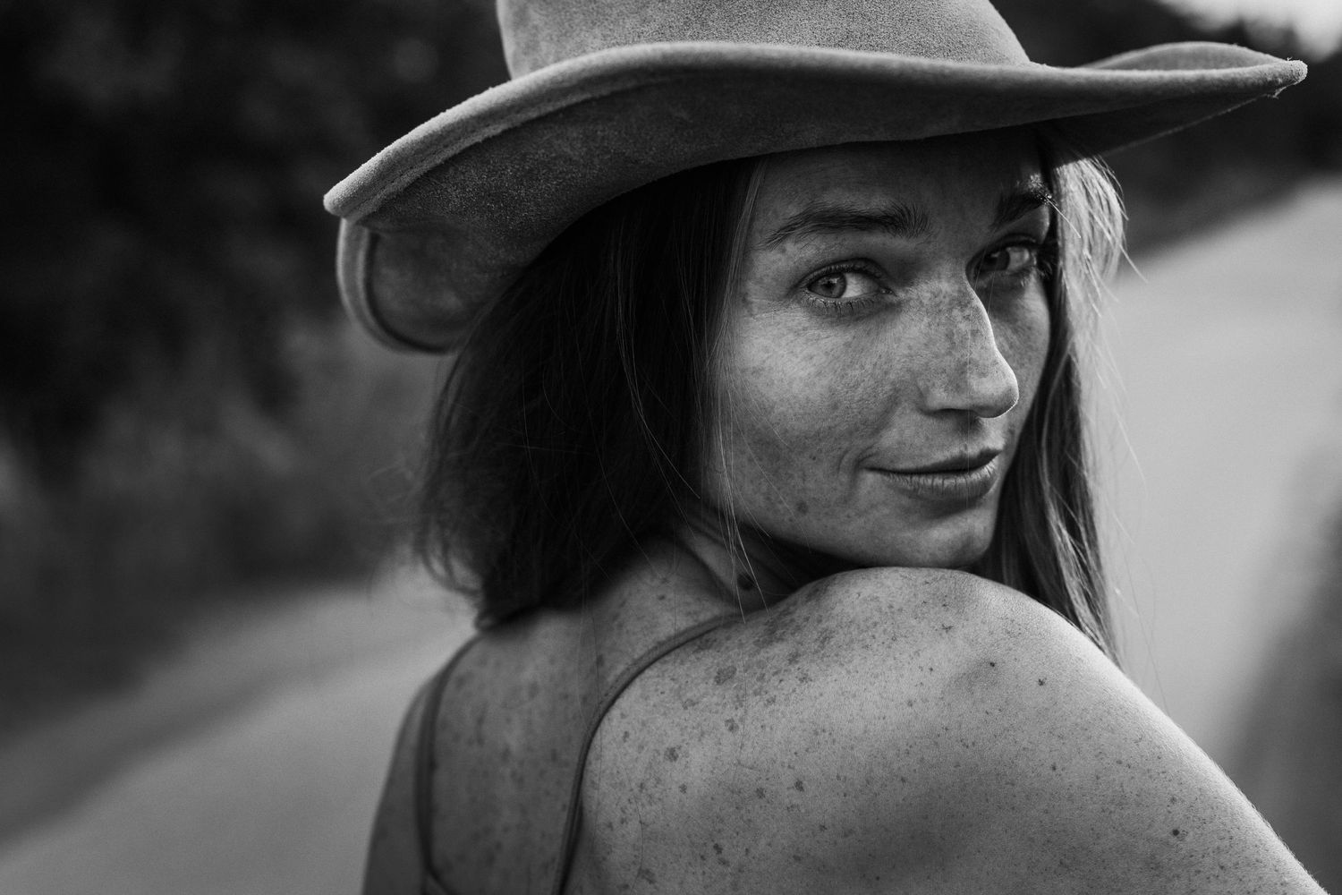 A black and white close-up portrait of a girl with a hat and freckles, looking over her shoulder and smiling.