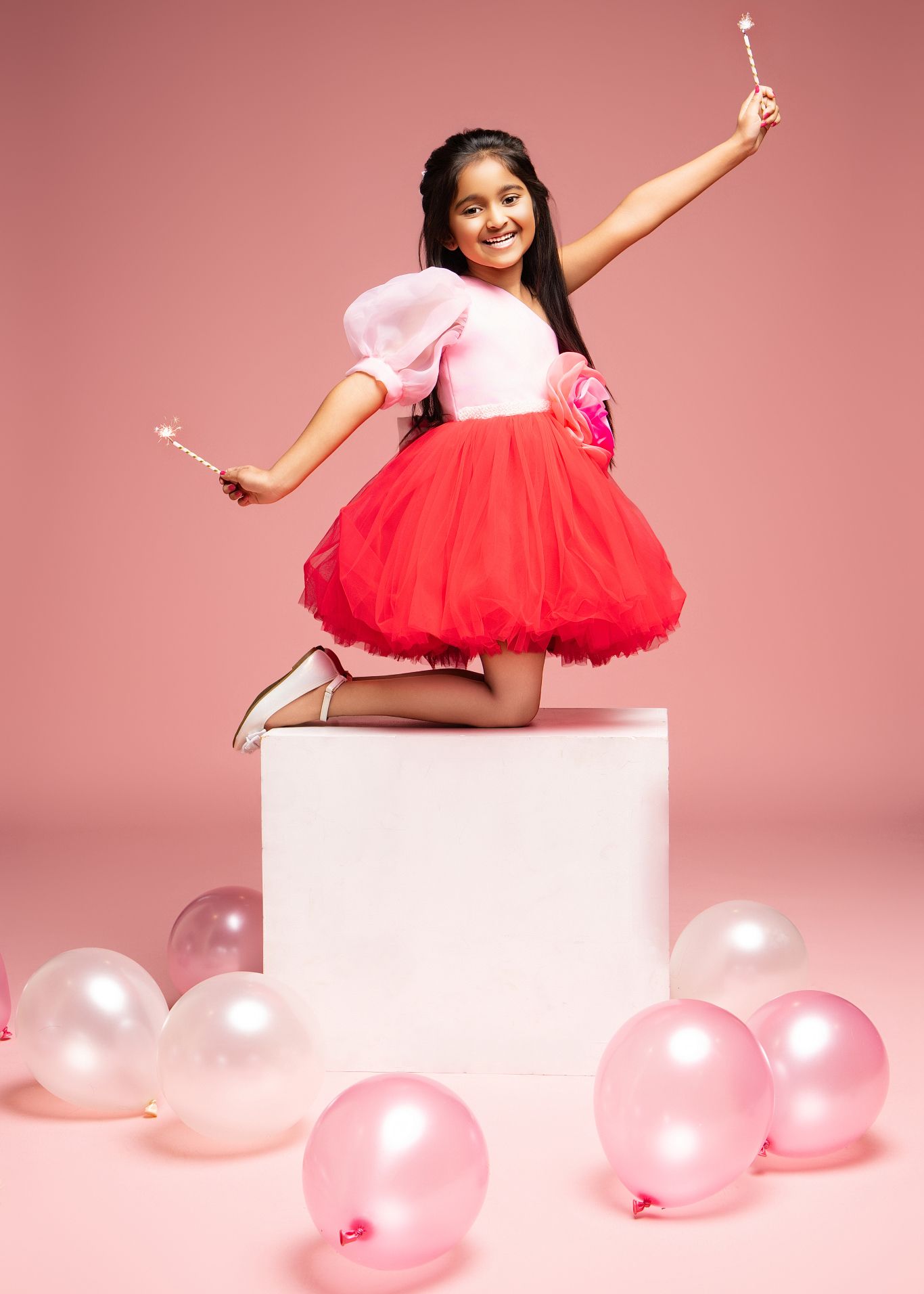 A person in a pink and red tulle dress poses playfully on a white platform surrounded by pink balloons against a rosy backdrop.