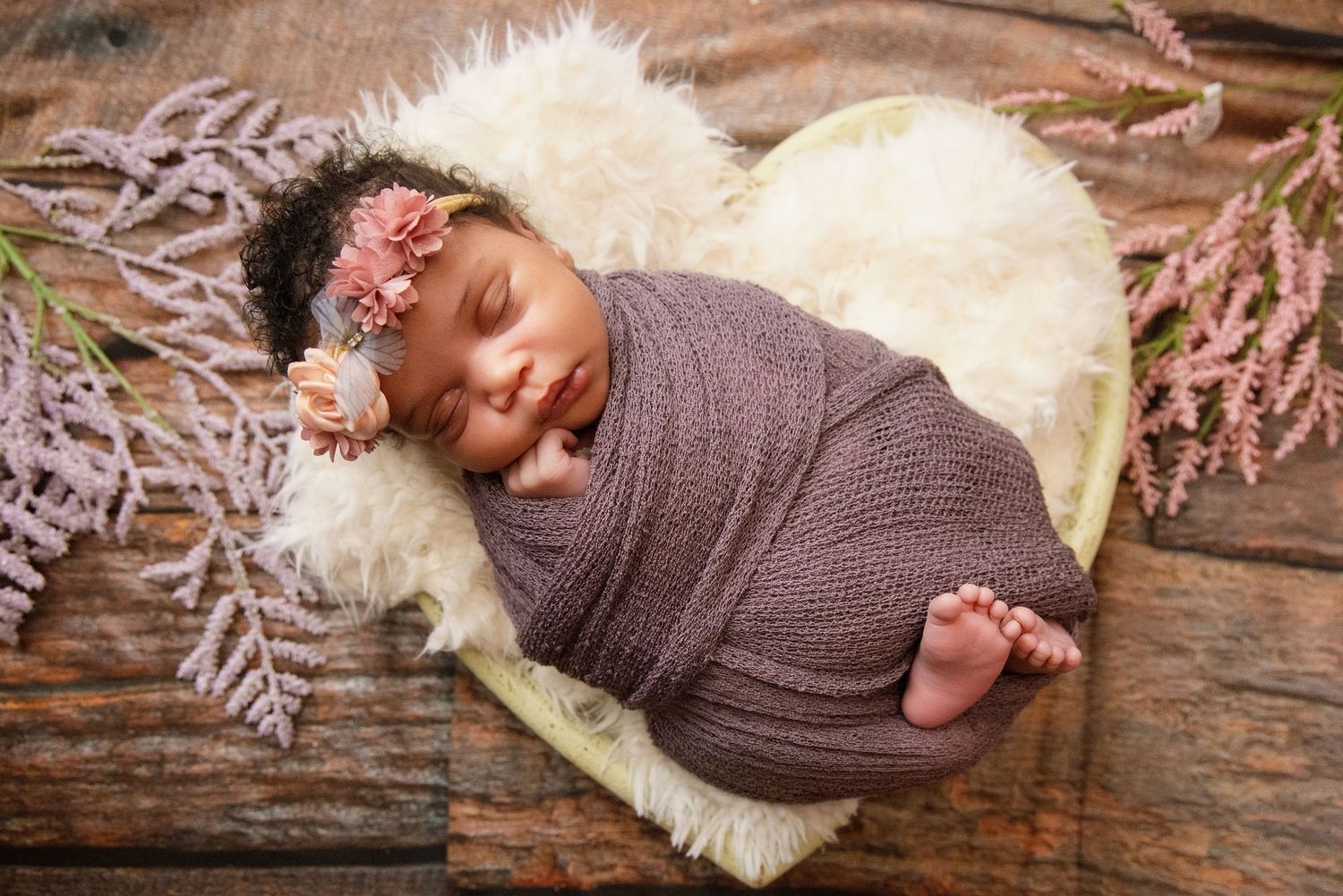 Newborn baby girl swaddled in mauve wrap, sleeping on heart-shaped fur prop with floral headband and lavender flowers.