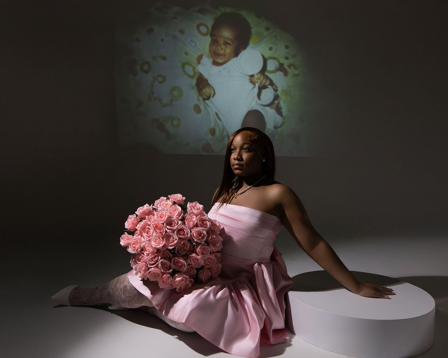 Woman in pink dress holding pink roses sits before projected childhood photo backdrop.
