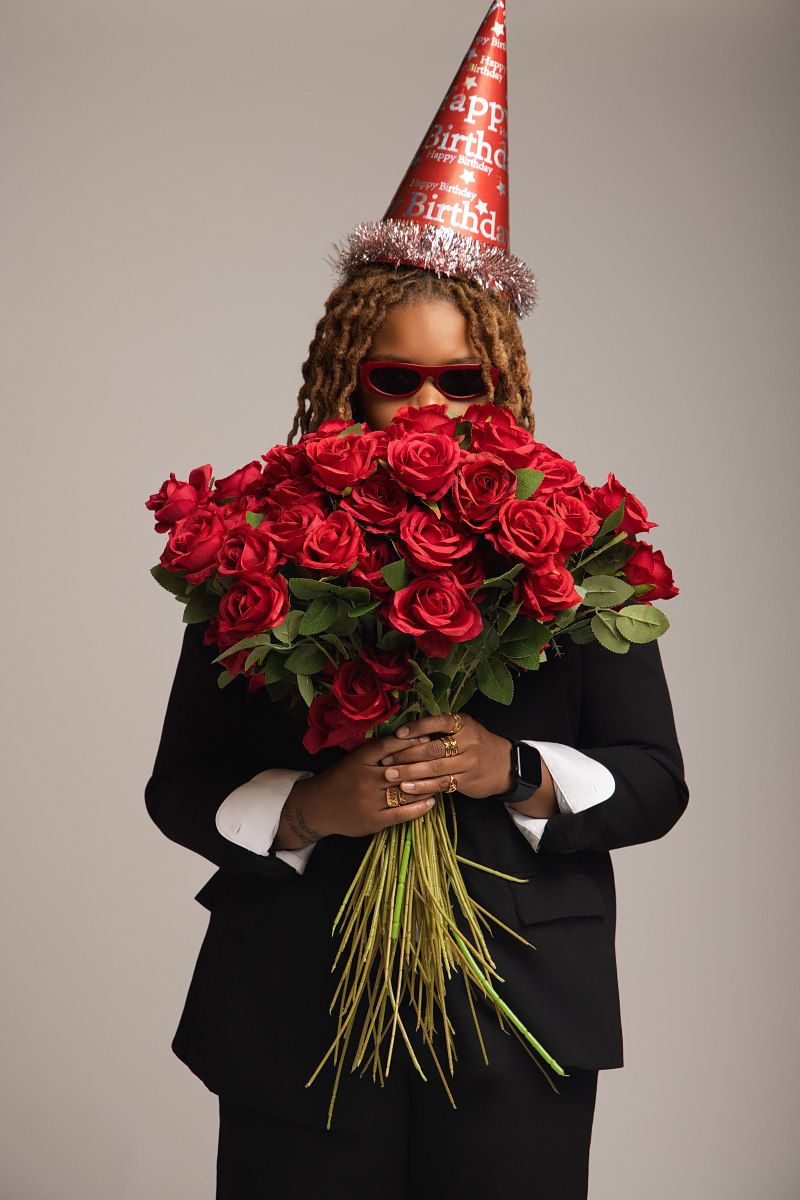 A figure in a black suit wearing a party hat holds a vibrant bouquet of red roses against a gray background.