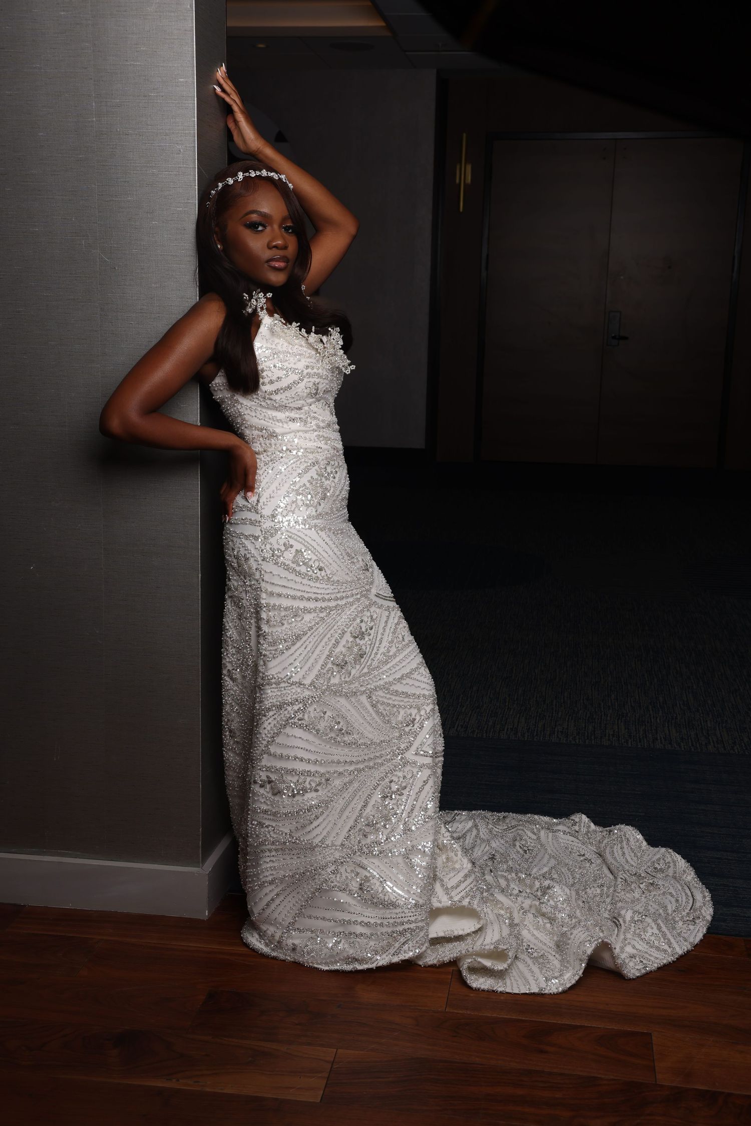 Elegant bride in an ornate white lace wedding gown with a long train poses dramatically against a dark wall.