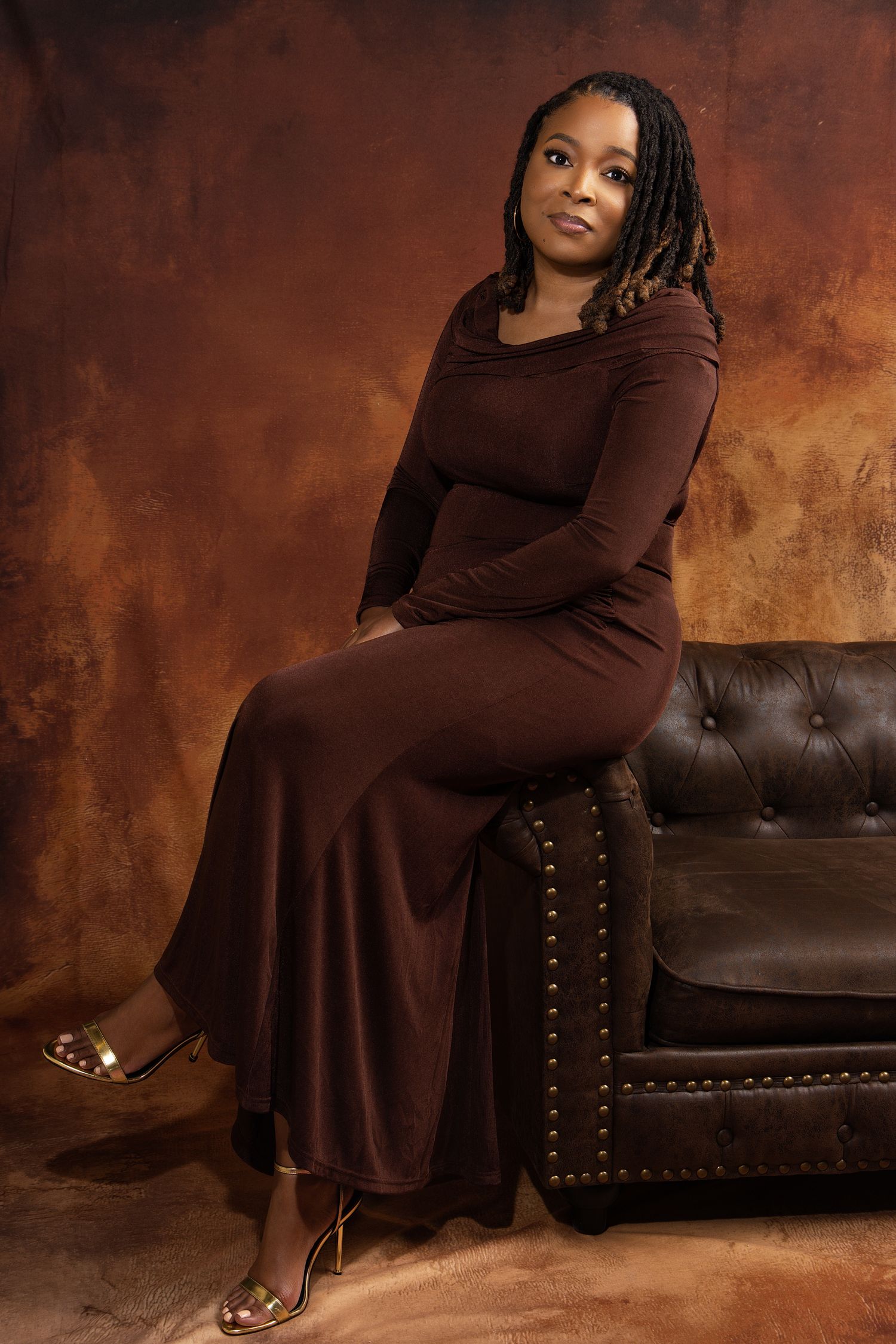 A woman in a brown long dress poses elegantly on a leather armchair against a textured brown backdrop.