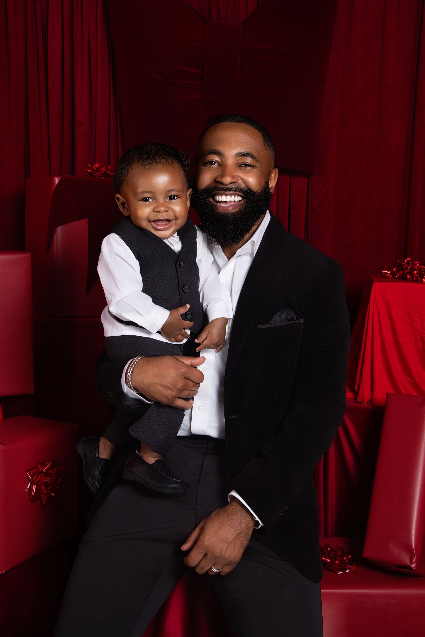 A parent and child share a joyful embrace while wearing formal black and white attire against a red backdrop.