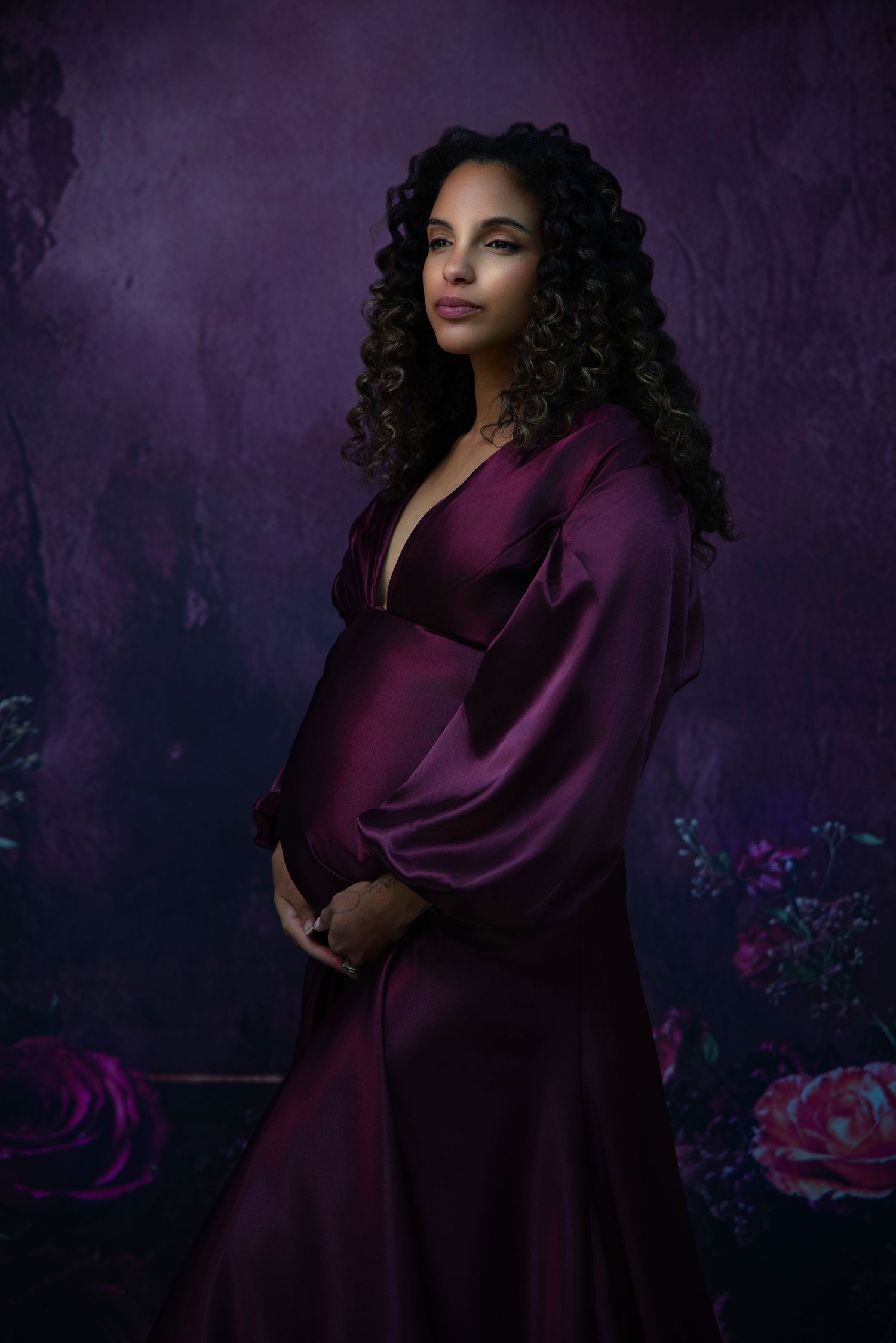 Artistic maternity photoshoot featuring a burgundy gown against a moody purple floral backdrop.