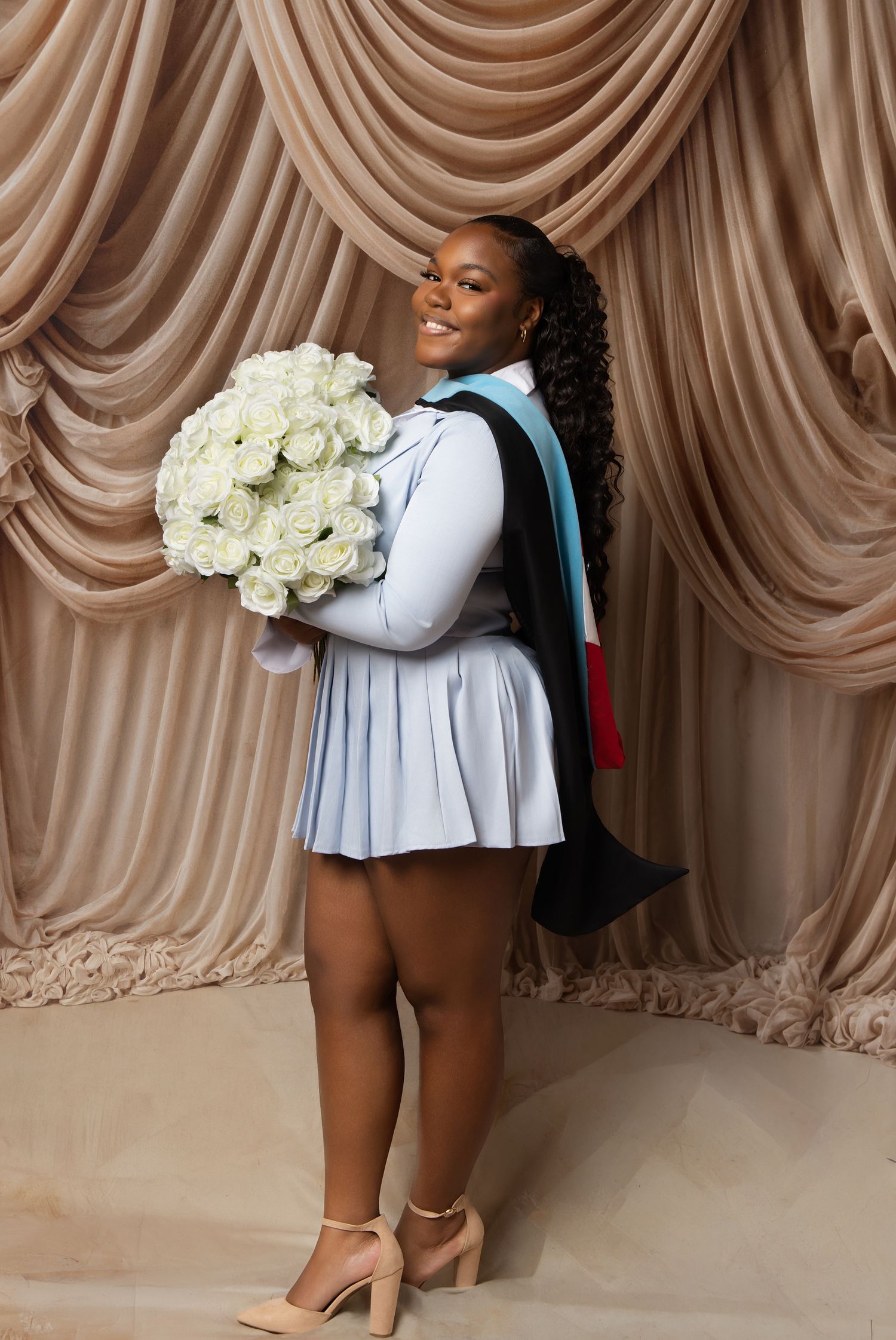 Smiling Black woman in graduation stole holding white roses bouquet wearing light blue dress and heels.