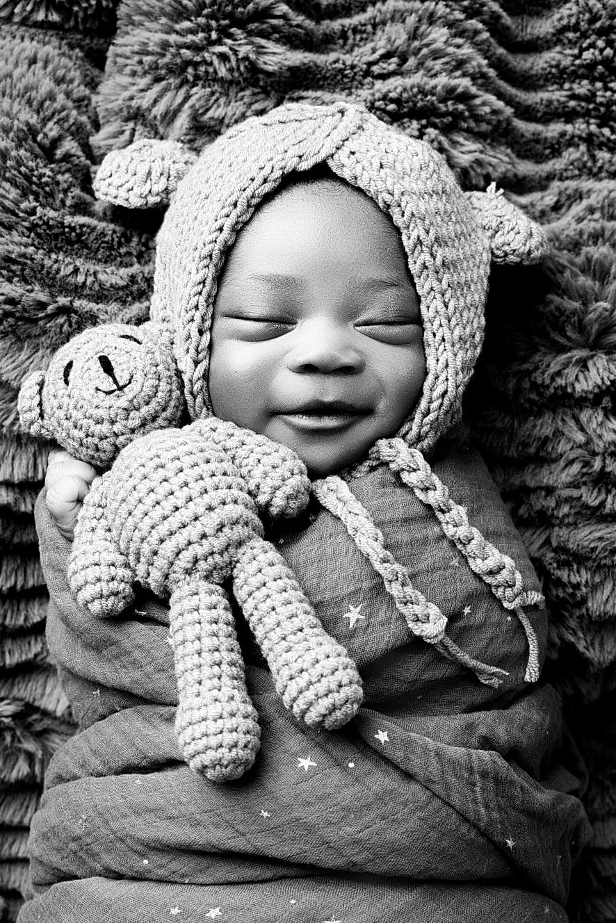 A joyful baby wrapped in a blanket wearing a knitted animal hat and holding a matching crocheted teddy bear toy.