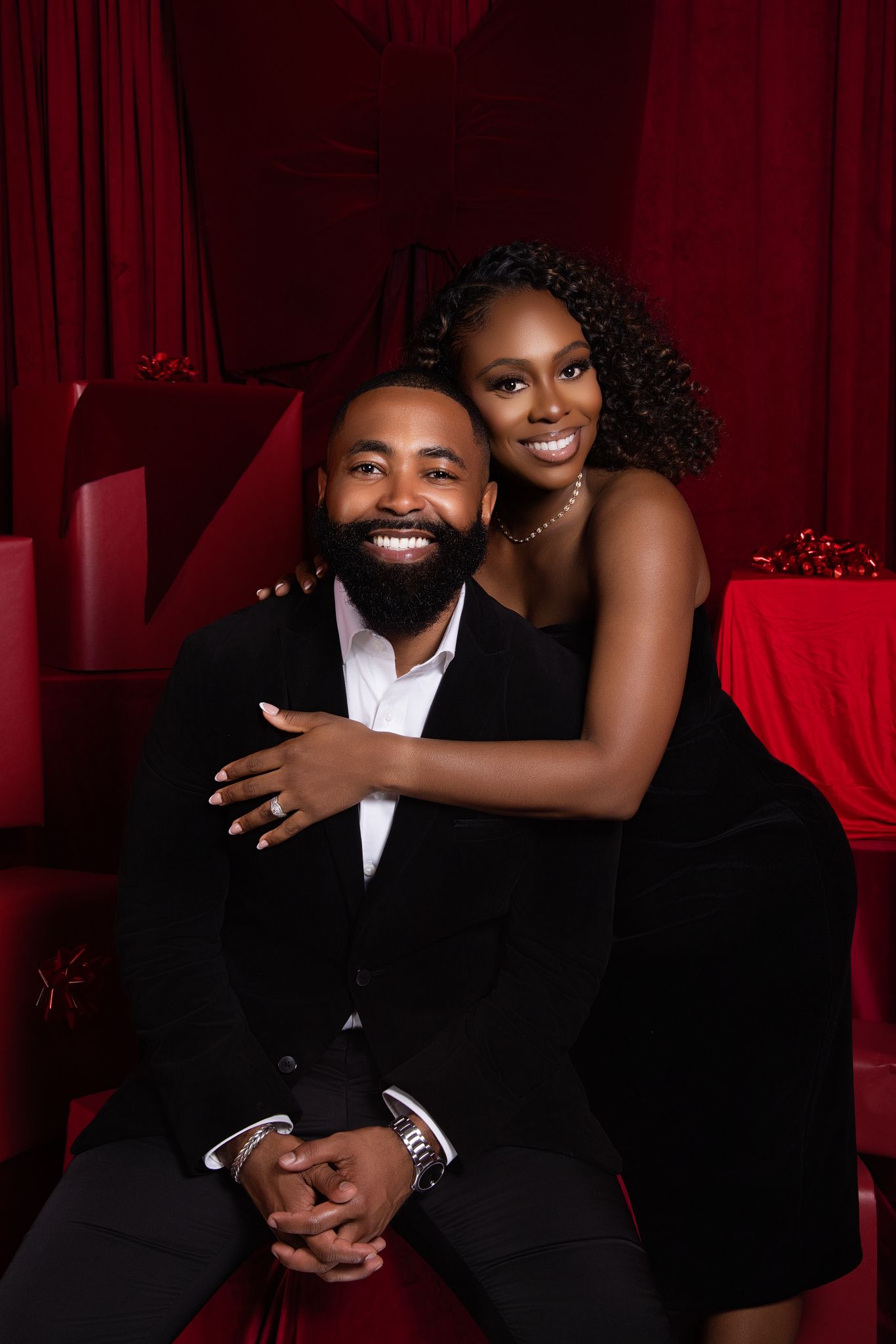 Romantic couple in formal attire pose intimately against red curtain backdrop during elegant photo session.