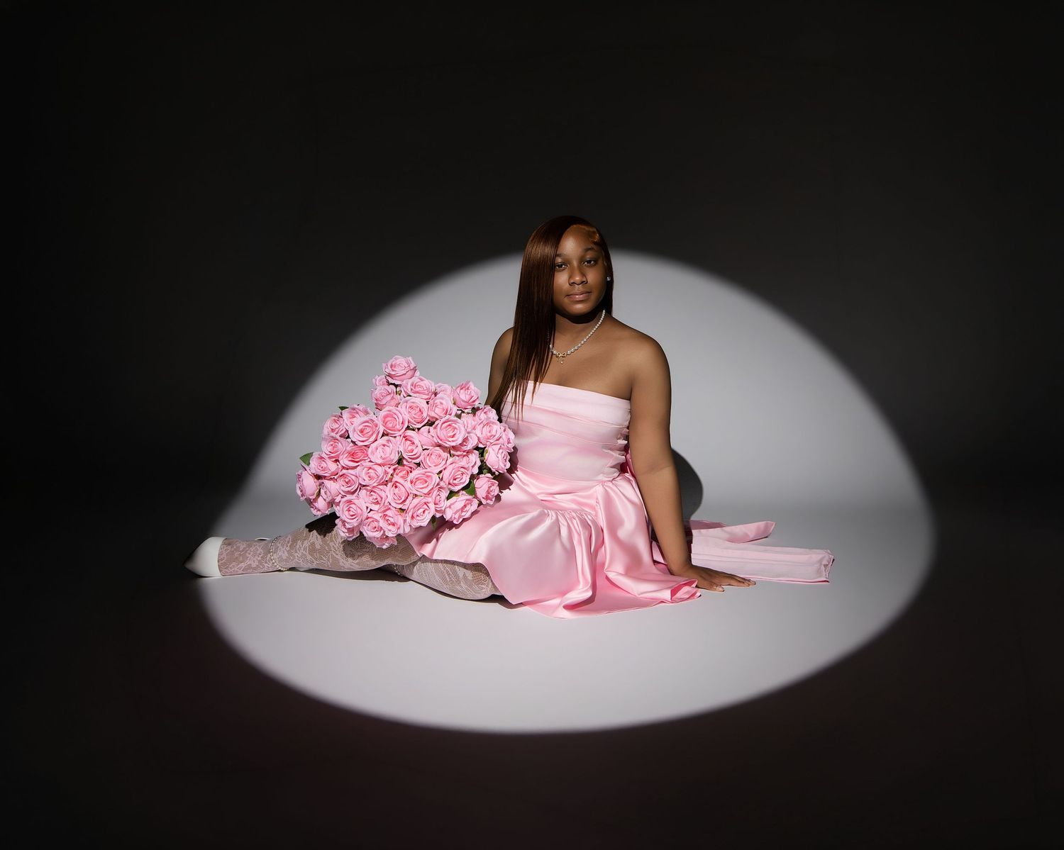 Woman in pink satin dress sitting in spotlight holding large bouquet of pink roses on dark background.