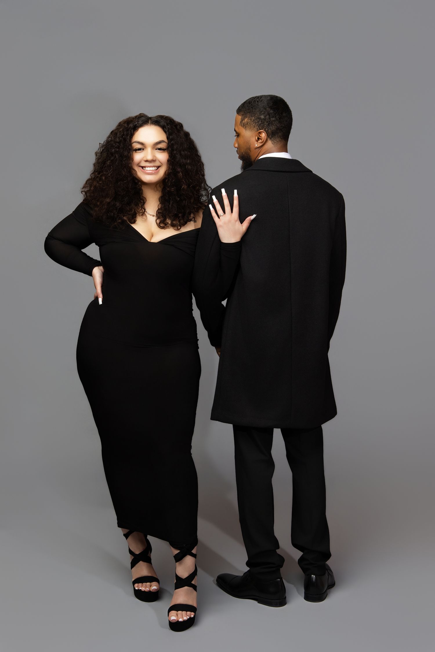 Two figures in black outfits pose together in a series of playful studio shots against a gray background.