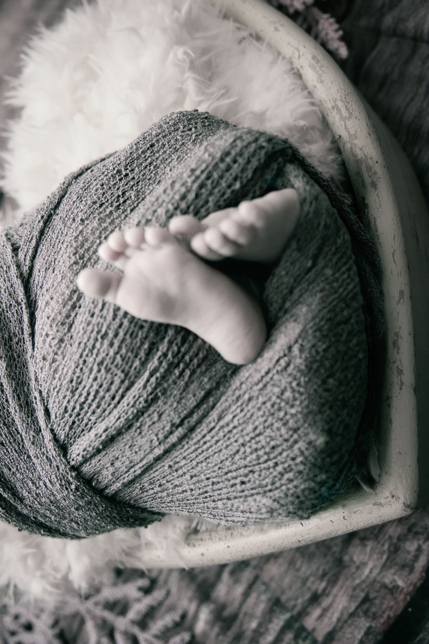 Black and white photo of newborn baby feet peeking out from a soft knit wrap in a wooden bowl.