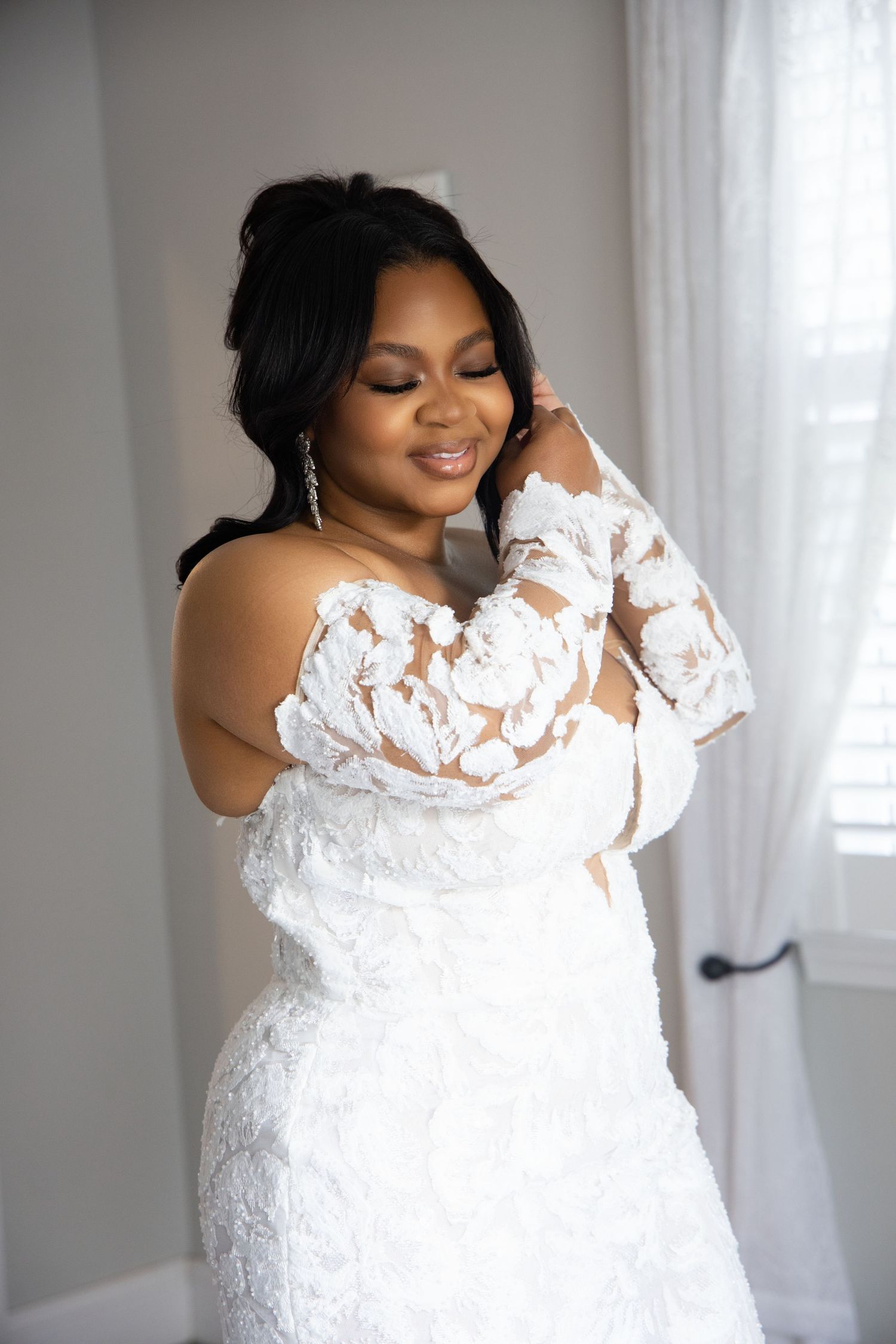 A bride wearing a stunning off-shoulder white lace wedding dress with long sleeves poses elegantly by a window.