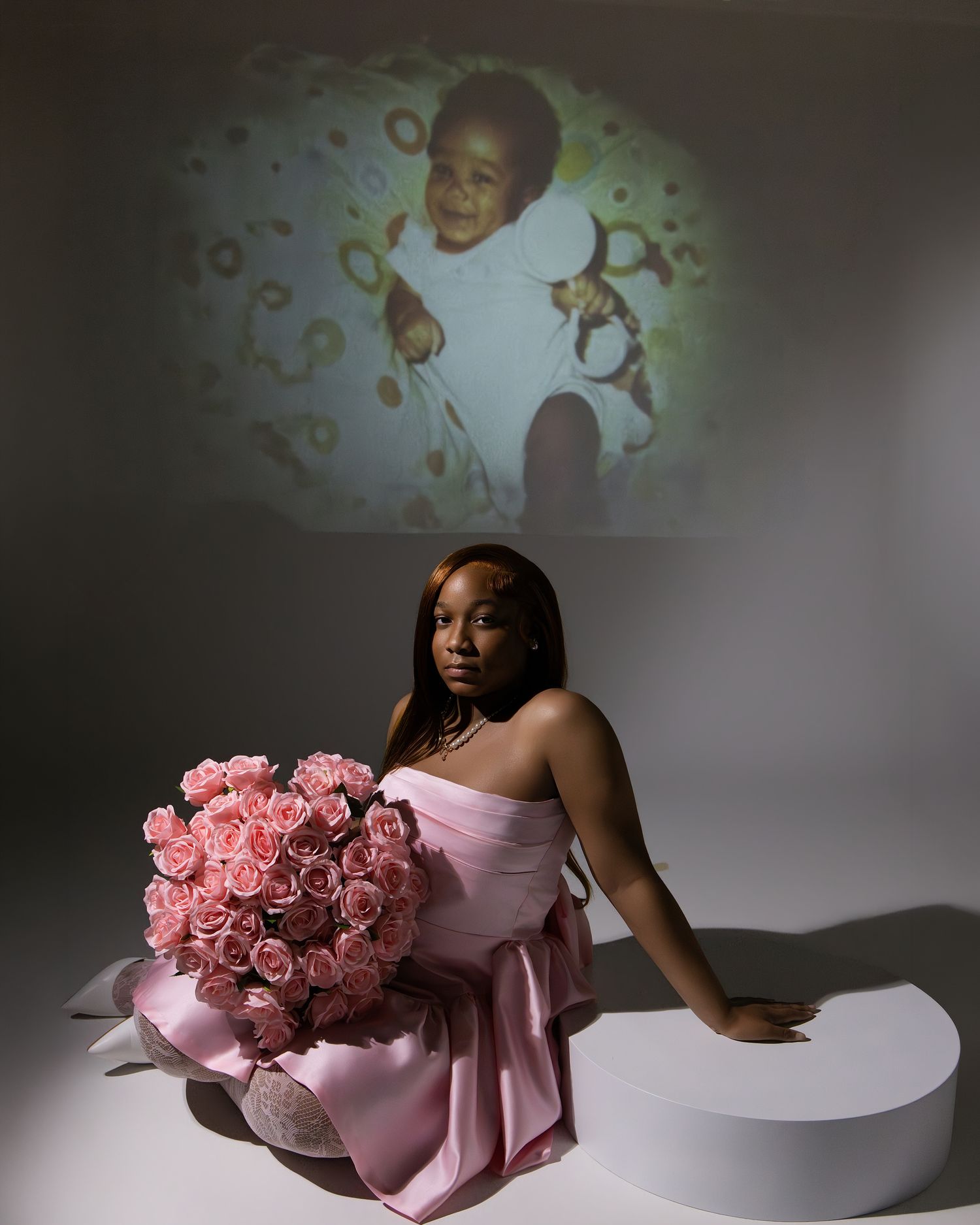 Woman in pink dress holds large pink rose bouquet, seated before projected childhood photo backdrop.