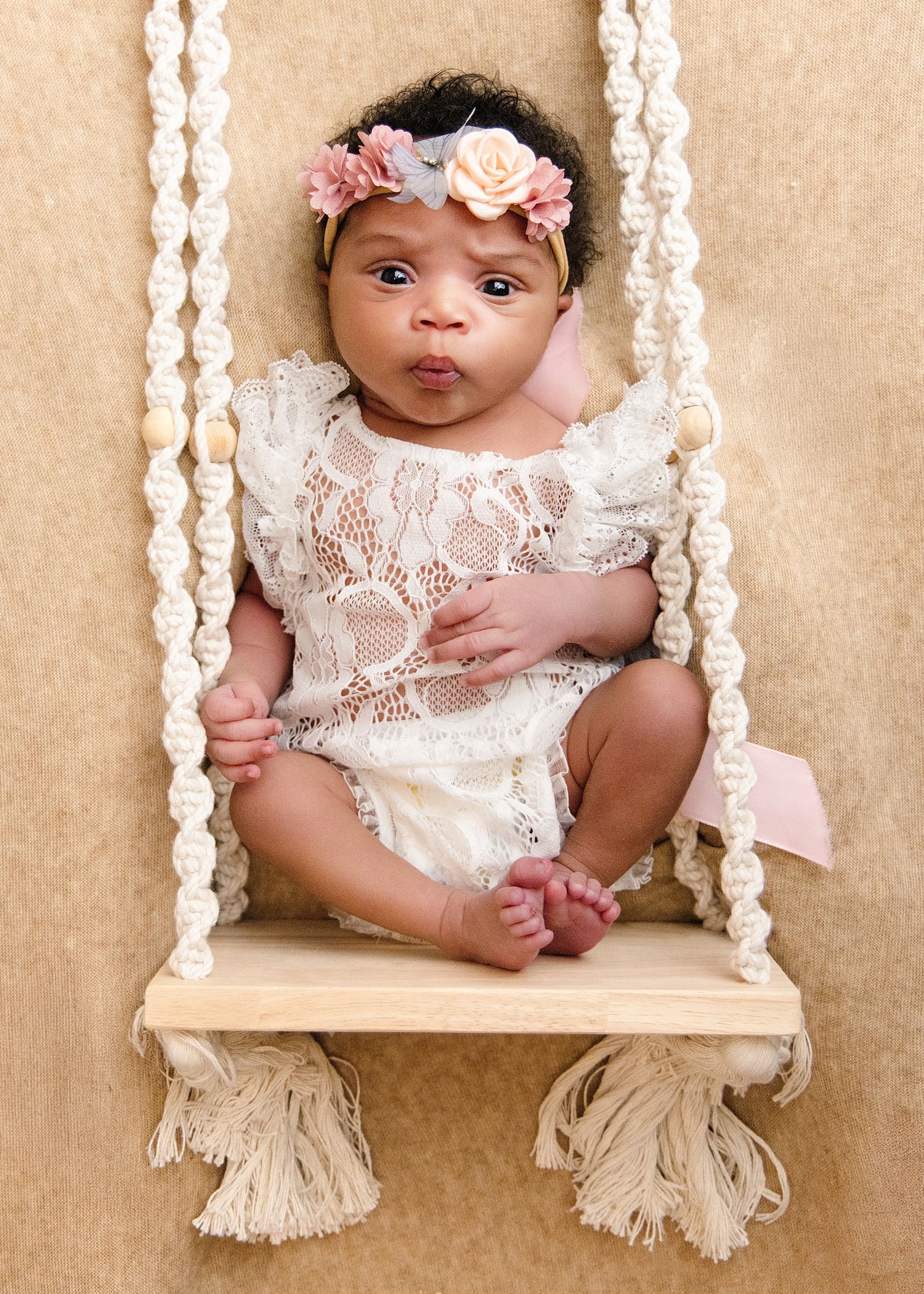 Newborn baby girl in white lace romper and floral headband sitting on a macrame swing for a professional photo.