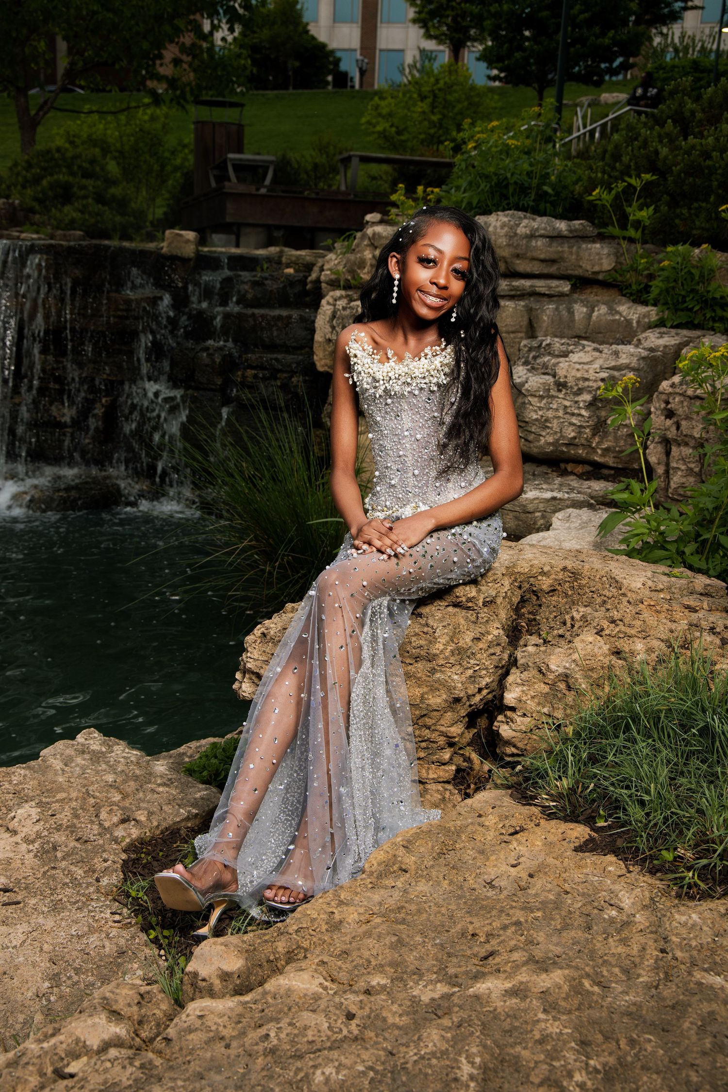 Woman in sparkly silver gown poses by waterfall on stone ledge.