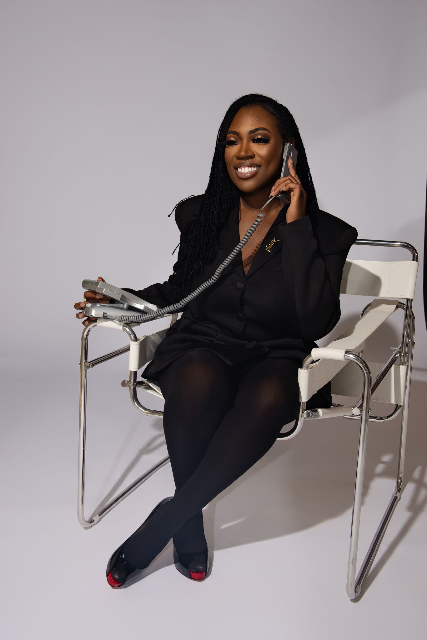 A person in a black outfit poses playfully on a modern white chair against a gray backdrop.