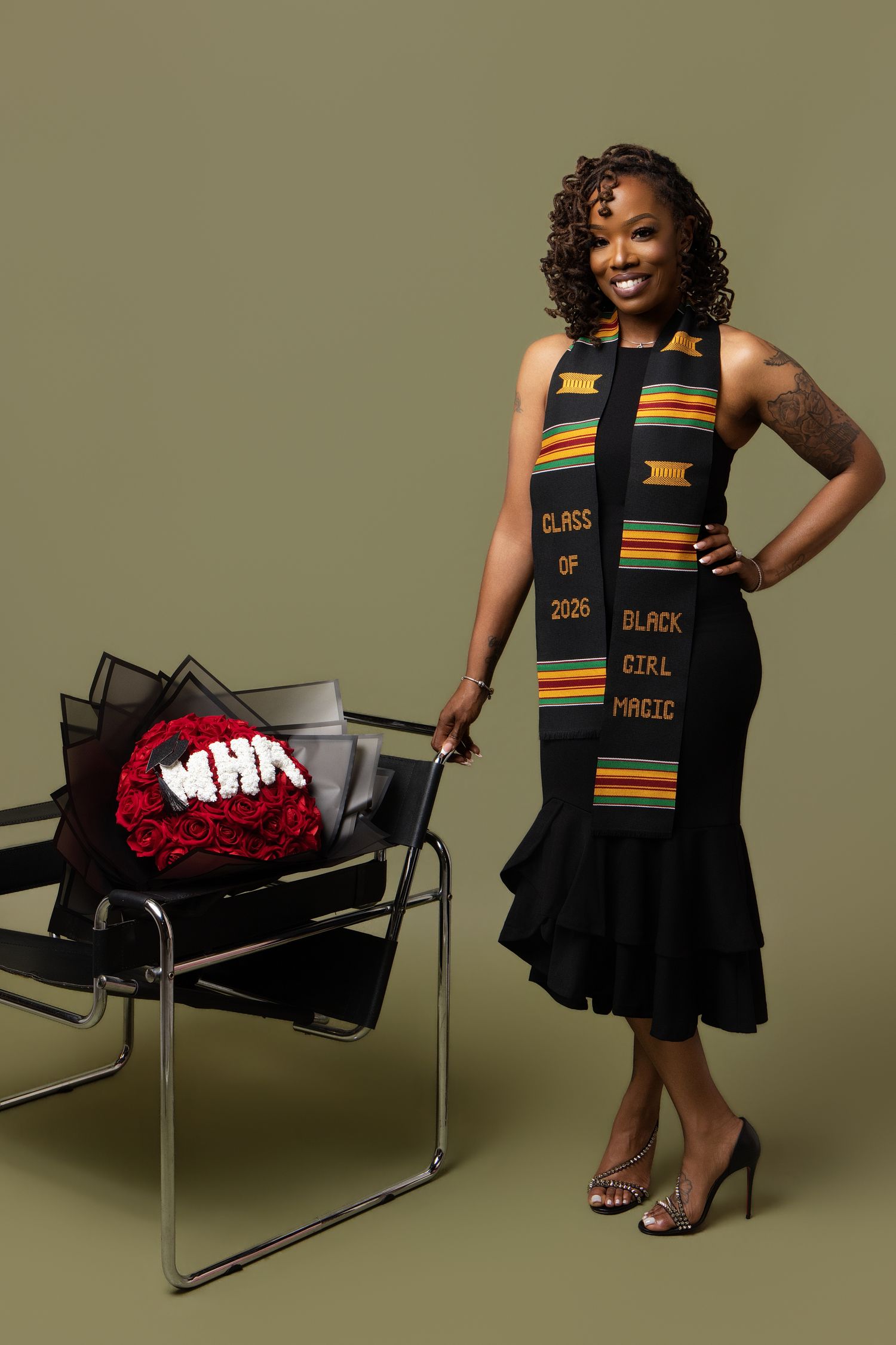 Woman in black dress wearing Class of 2026 Black Girl Magic kente graduation stole, posing beside chair with red roses.