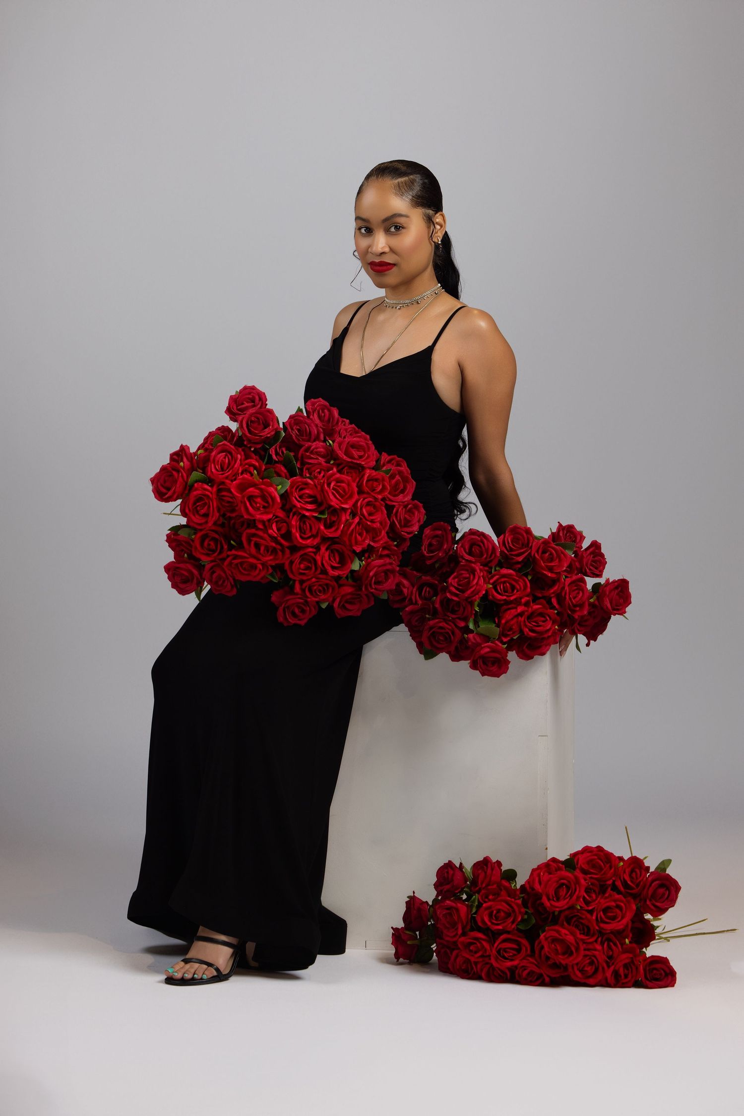 A woman in a black maxi dress poses elegantly with bouquets of vibrant red roses against a gray studio backdrop.