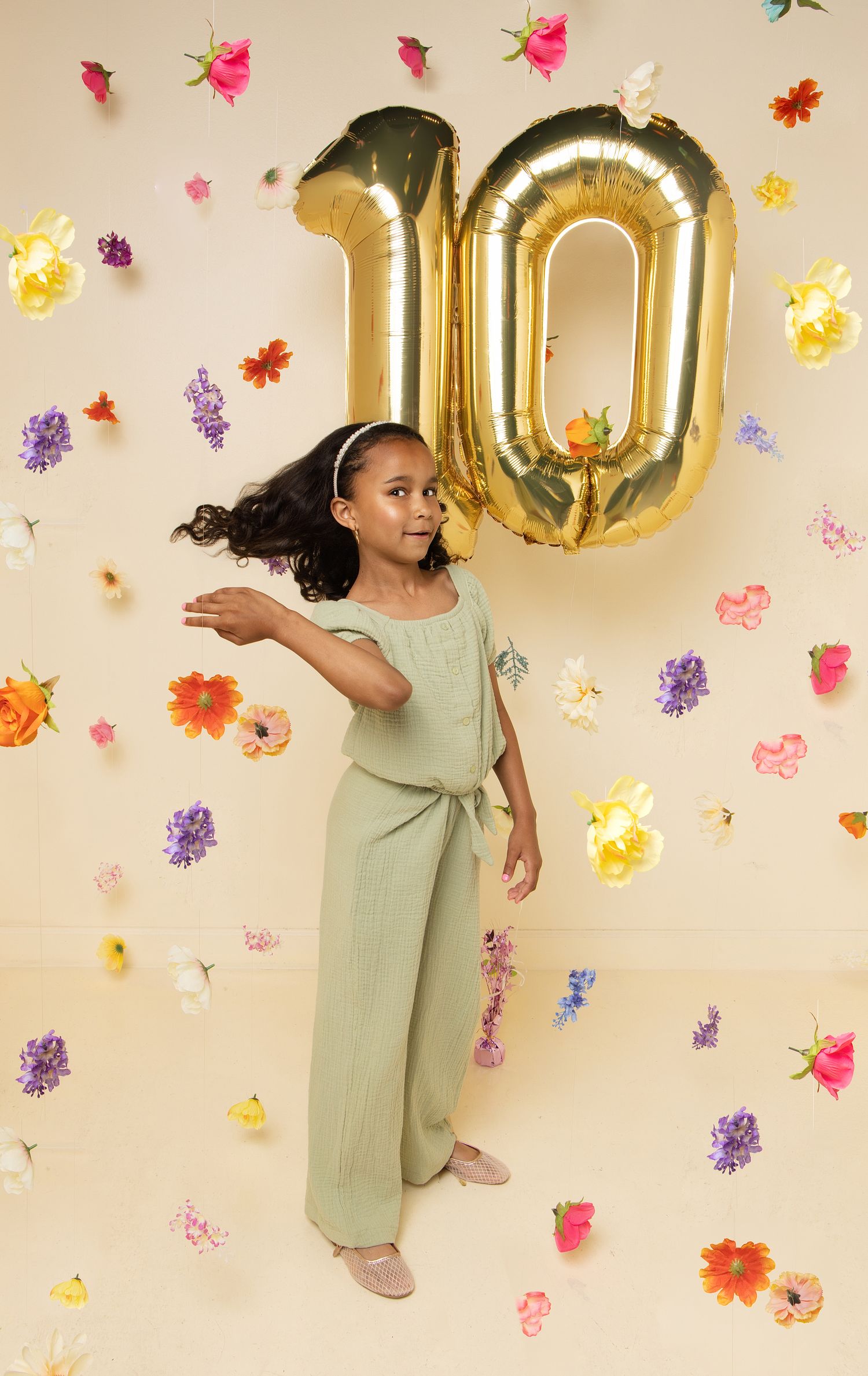 A young girl in a green jumpsuit poses joyfully in front of a floral backdrop with gold number 10 balloons.