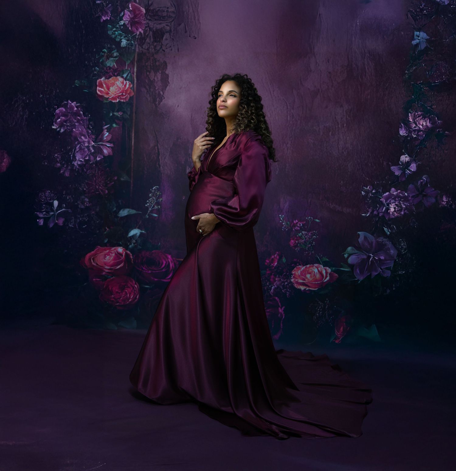Dramatic maternity photos in deep purple dress with artistic rose backdrop and moody lighting.