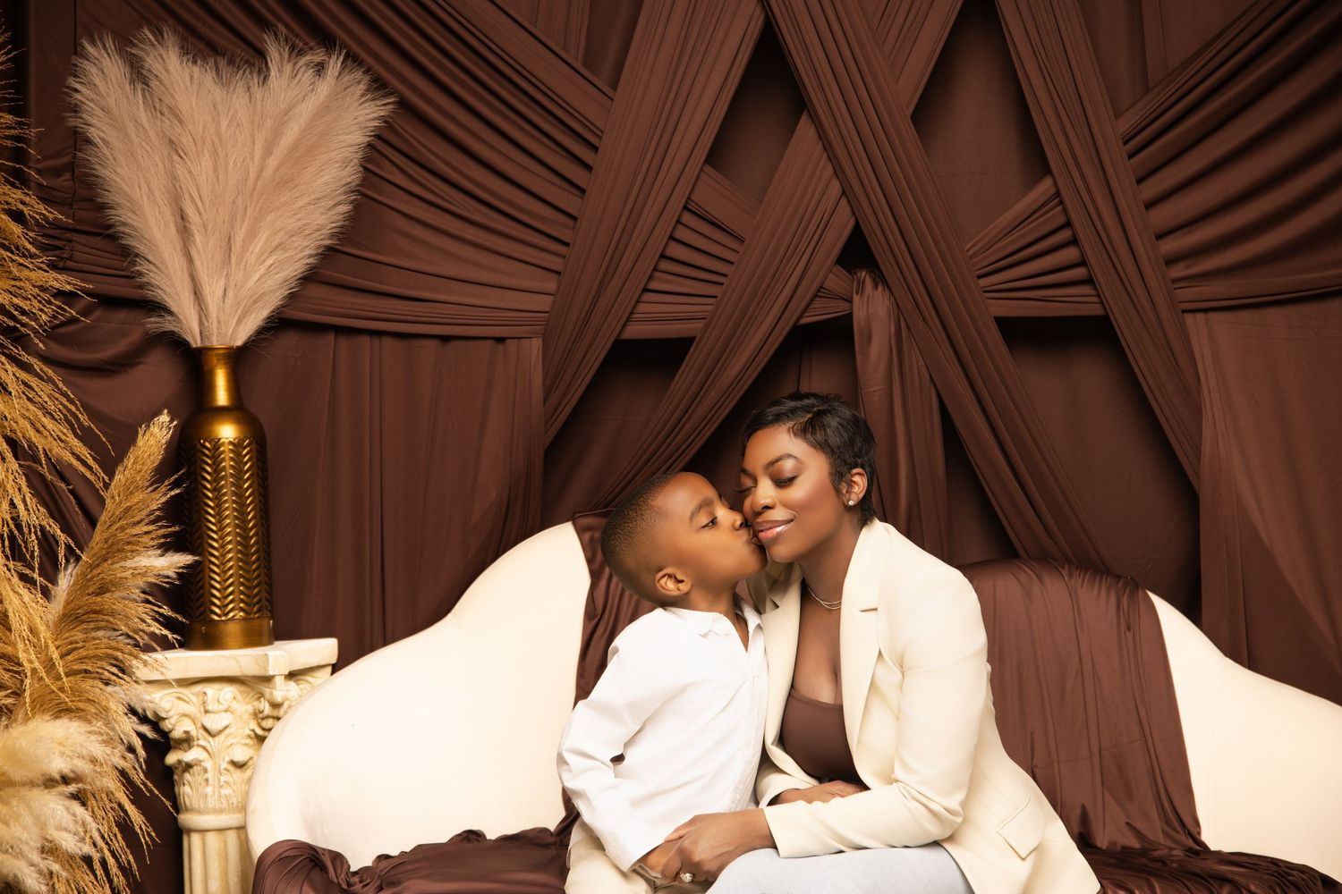A stylish couple in white outfits poses together against a brown draped backdrop with feather decorations.