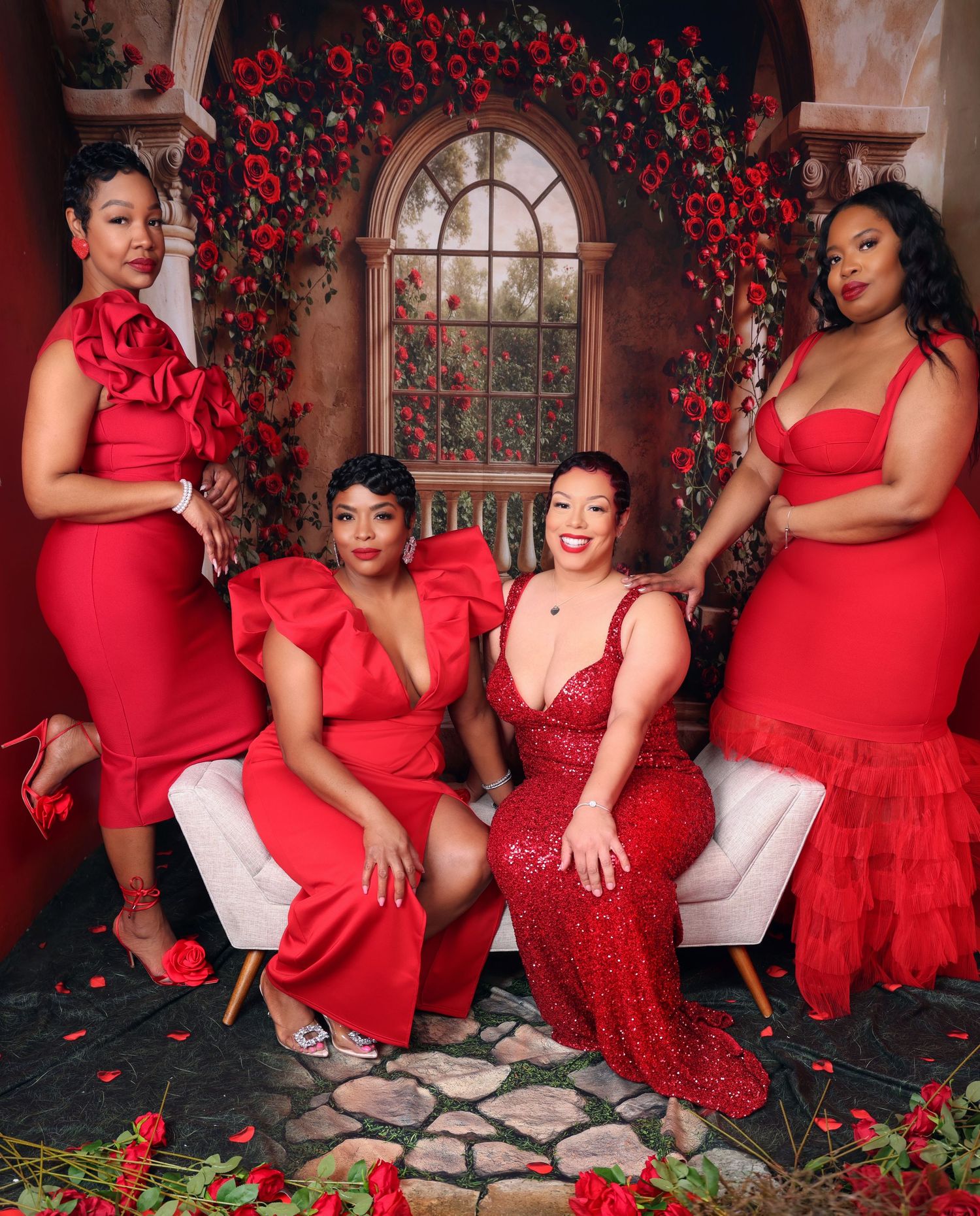 Four women in stunning red formal dresses pose beneath floral arch with roses.
