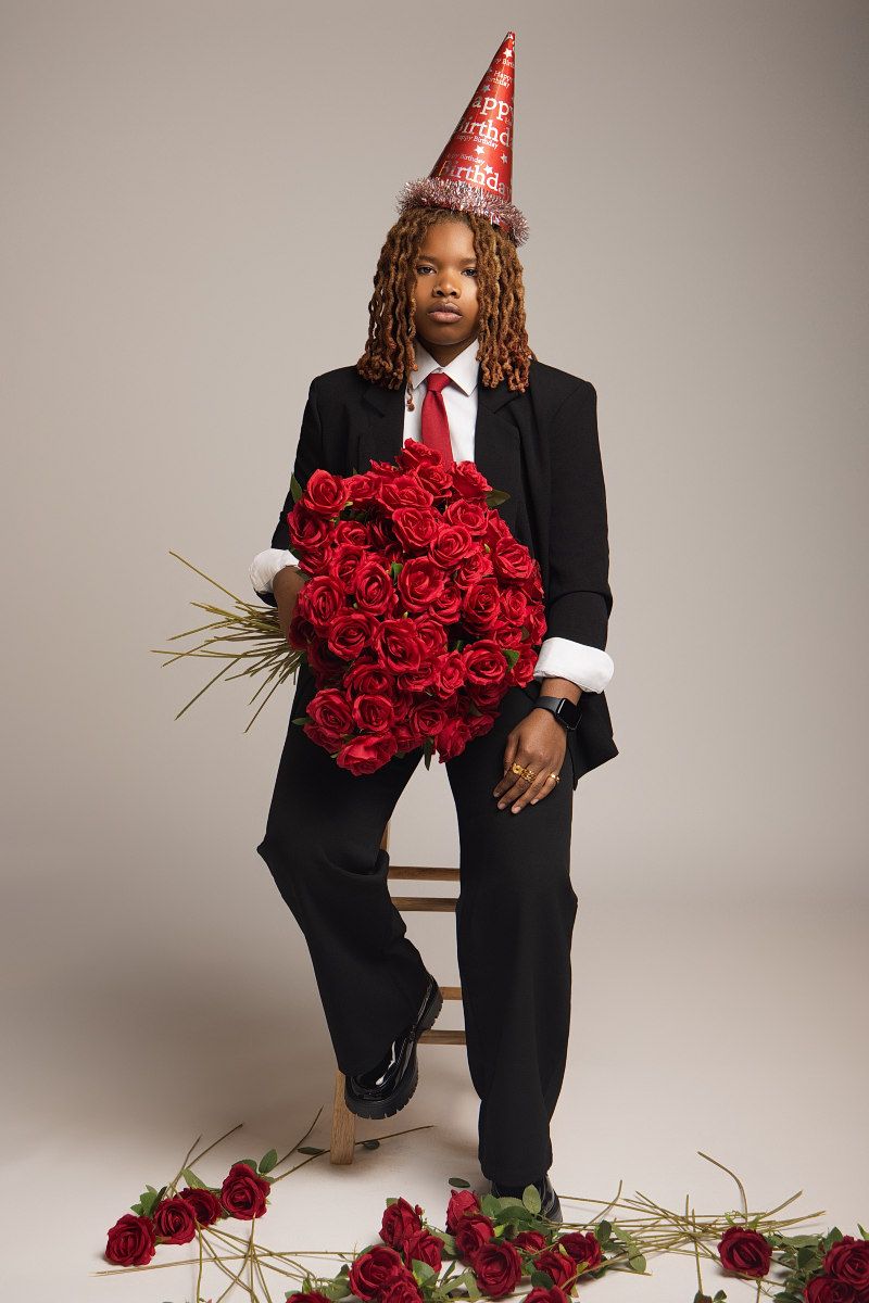 Someone in a black suit and party hat holds vibrant red roses against a neutral studio backdrop.