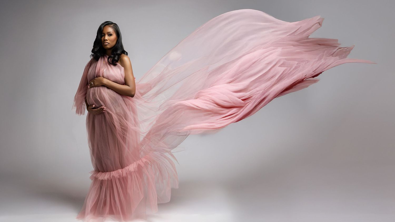 A pregnant woman in a flowing pink dress poses gracefully against a gray backdrop in a maternity photoshoot sequence.