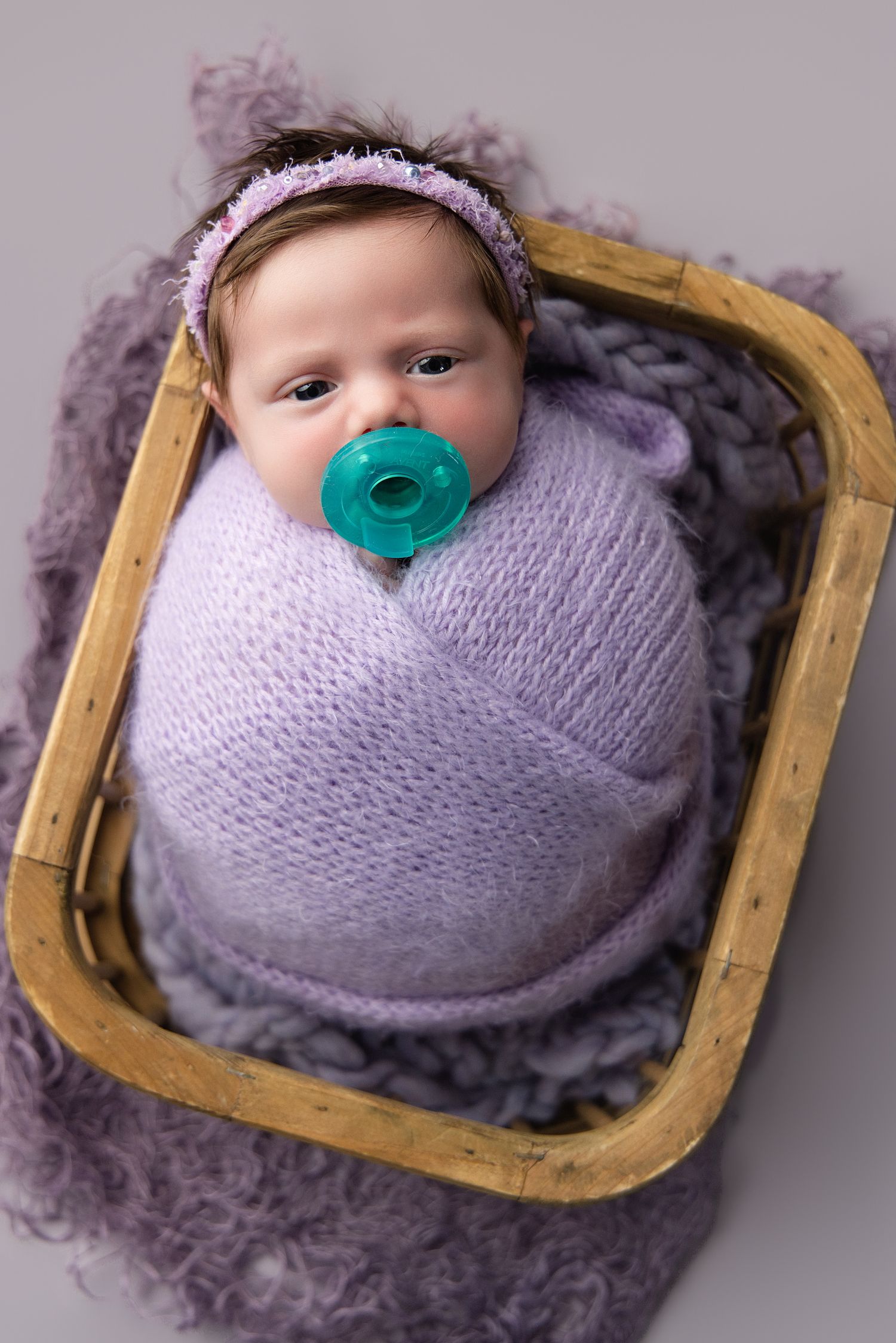 Newborn baby girl swaddled in purple wrap with teal pacifier, resting in wooden basket on lavender fluffy blanket.