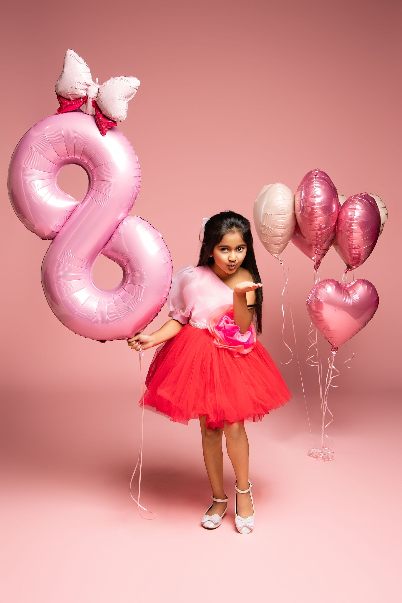 Pink balloon numbers create a whimsical backdrop for a festive photoshoot with a red tulle dress against a rosy background.