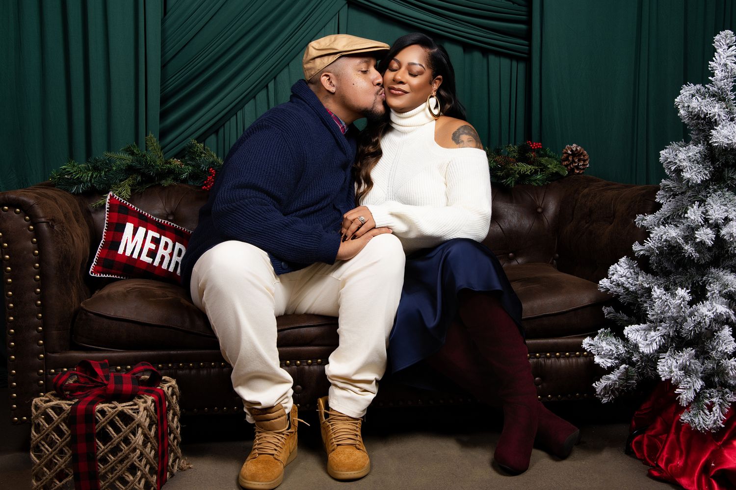 A couple sits on a wooden bench in front of a dark green backdrop with winter holiday decorations.