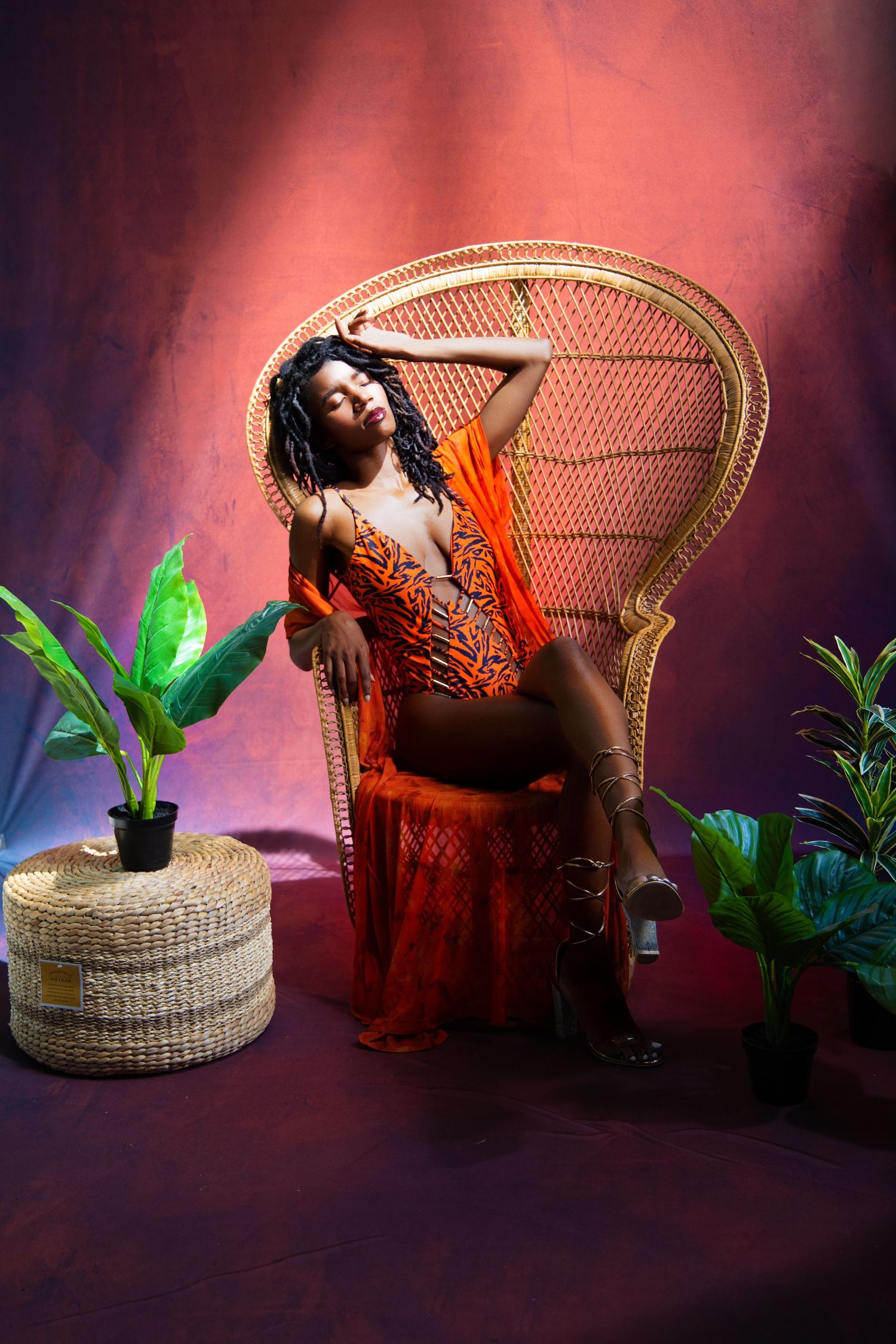 Stylish portrait session with wicker peacock chair and orange dress against red backdrop.