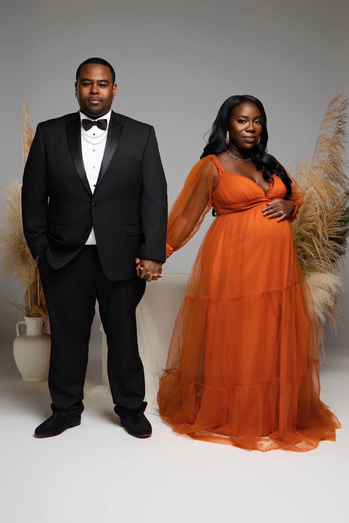 A couple poses in formal attire with a black suit and flowing burnt orange gown against a gray studio backdrop.