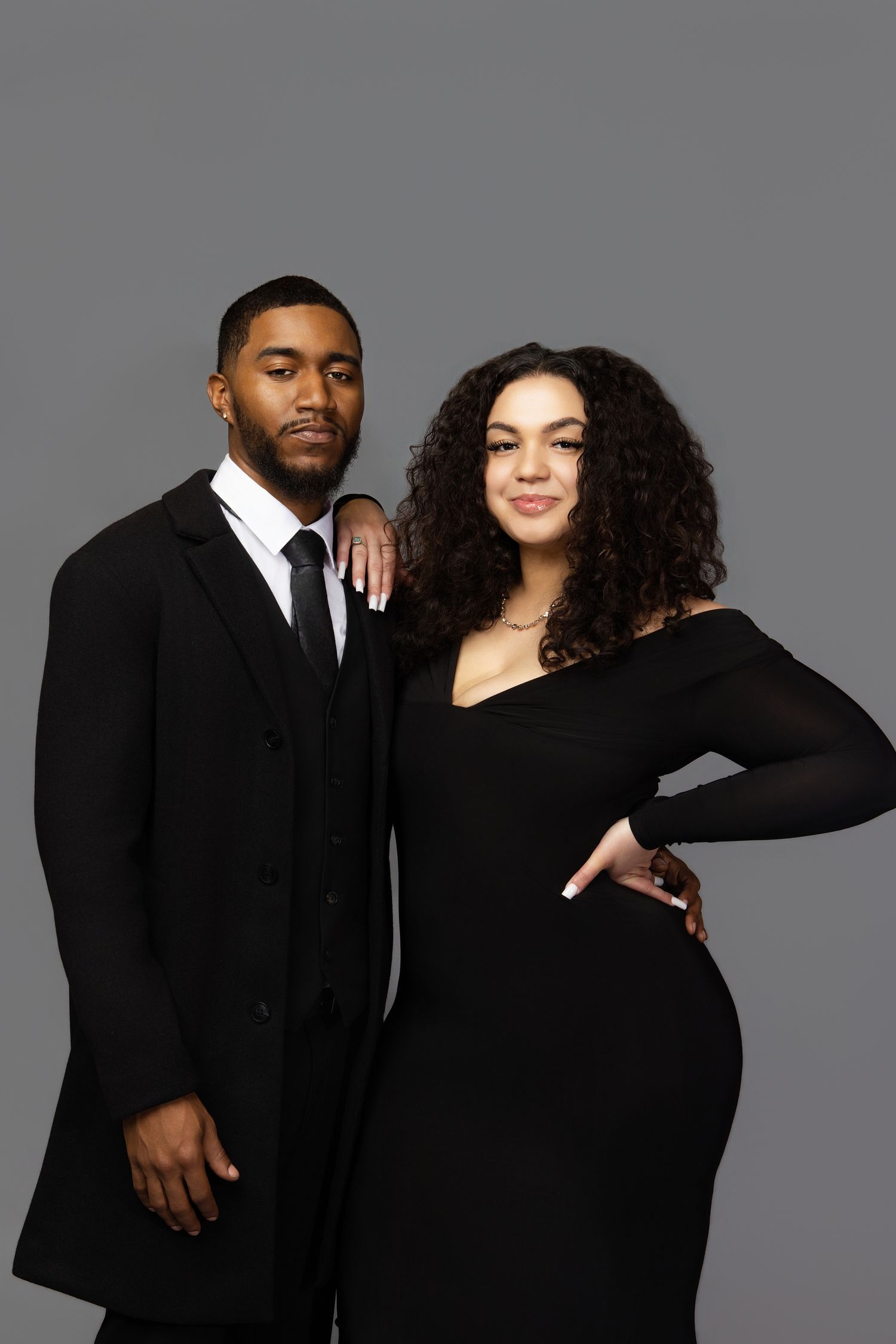A couple in matching black outfits poses together against a gray studio backdrop in a series of intimate portraits.
