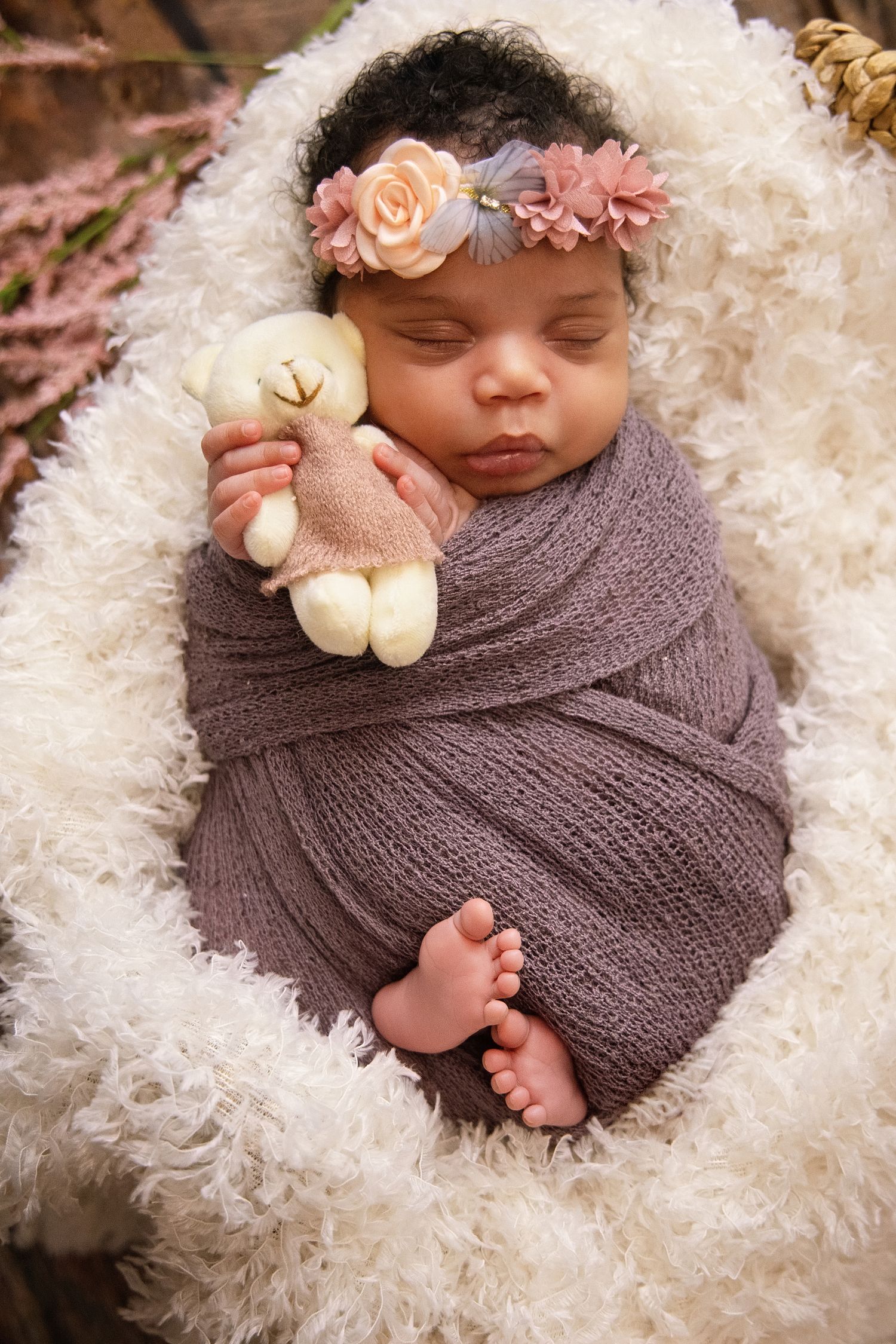 Newborn baby swaddled in purple wrap holding teddy bear, wearing floral headband, sleeping peacefully on fluffy white blanket.