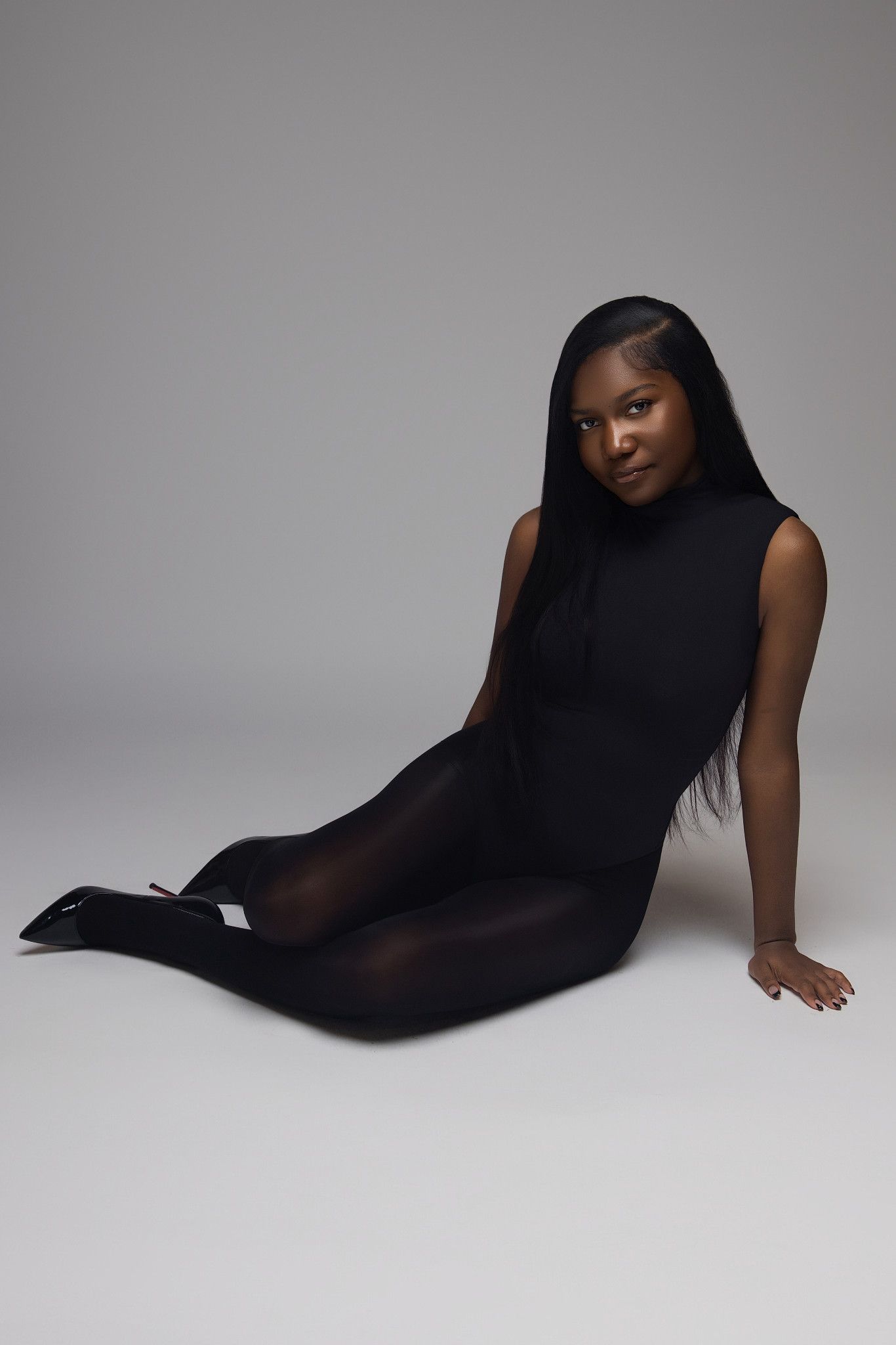 A figure in a black sleeveless bodysuit poses artistically against a light gray studio backdrop.