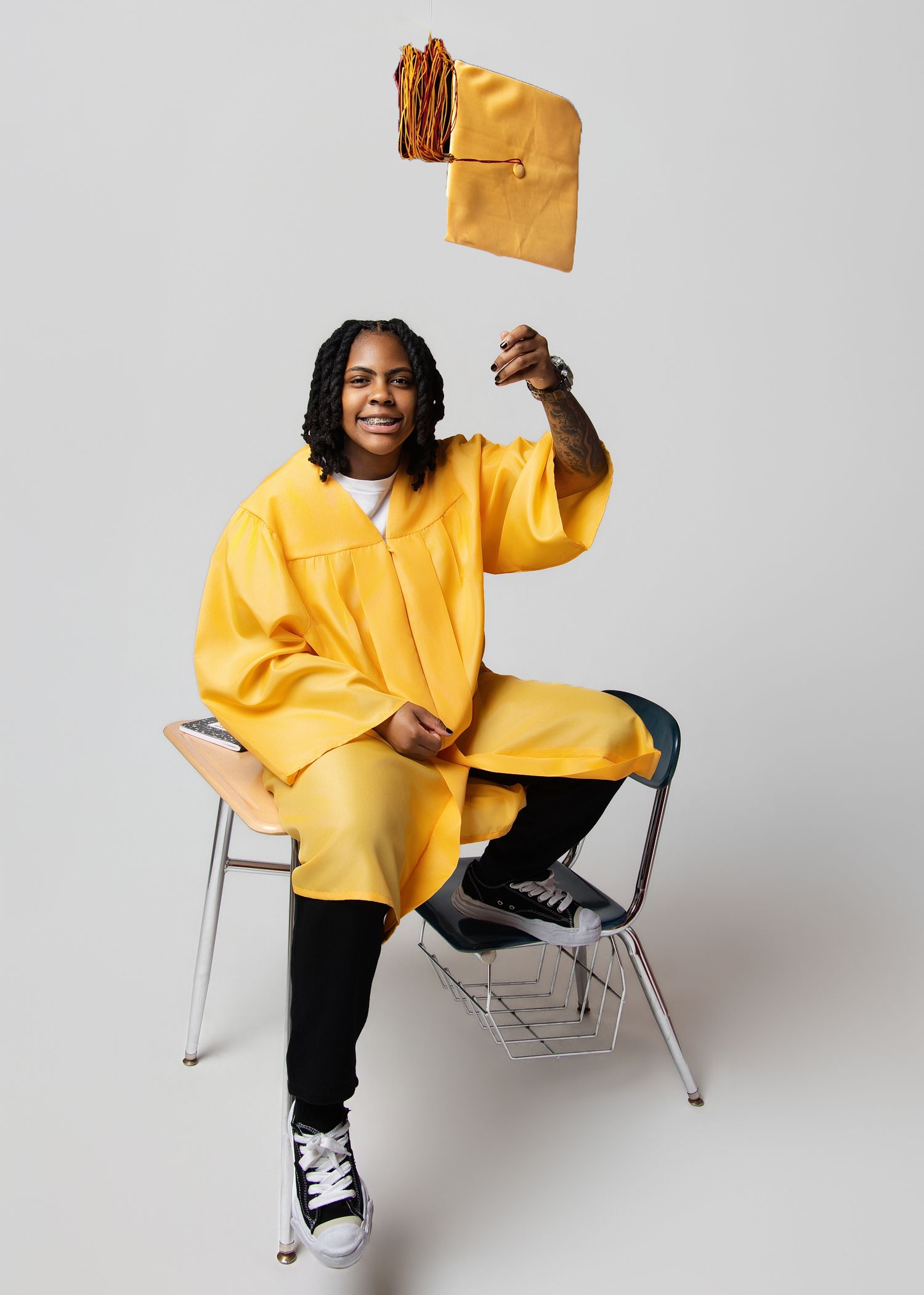A person in a bright yellow hoodie and black pants poses playfully on a modern chair against a white background.