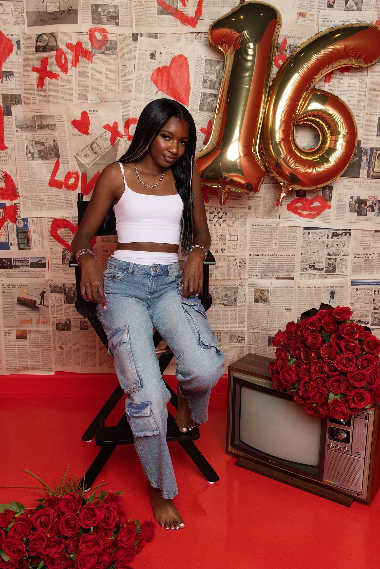 Young woman in white crop top and jeans poses on chair with gold number 16 balloon and red roses for sweet sixteen celebration.
