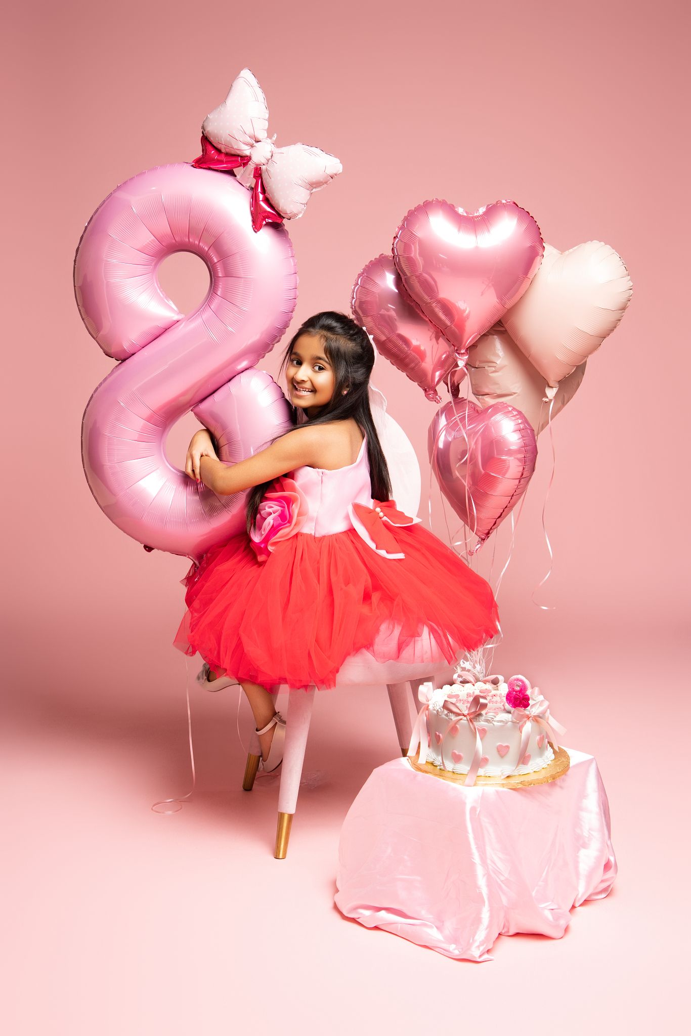 Pink balloon numbers and red tulle dress create a whimsical birthday photoshoot setup against a rosy backdrop.