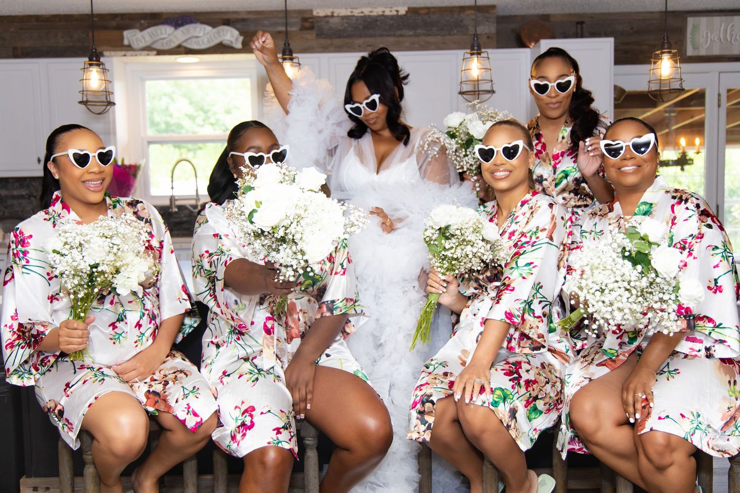 A bridal party wearing matching floral robes and sunglasses poses together while holding white bouquets.
