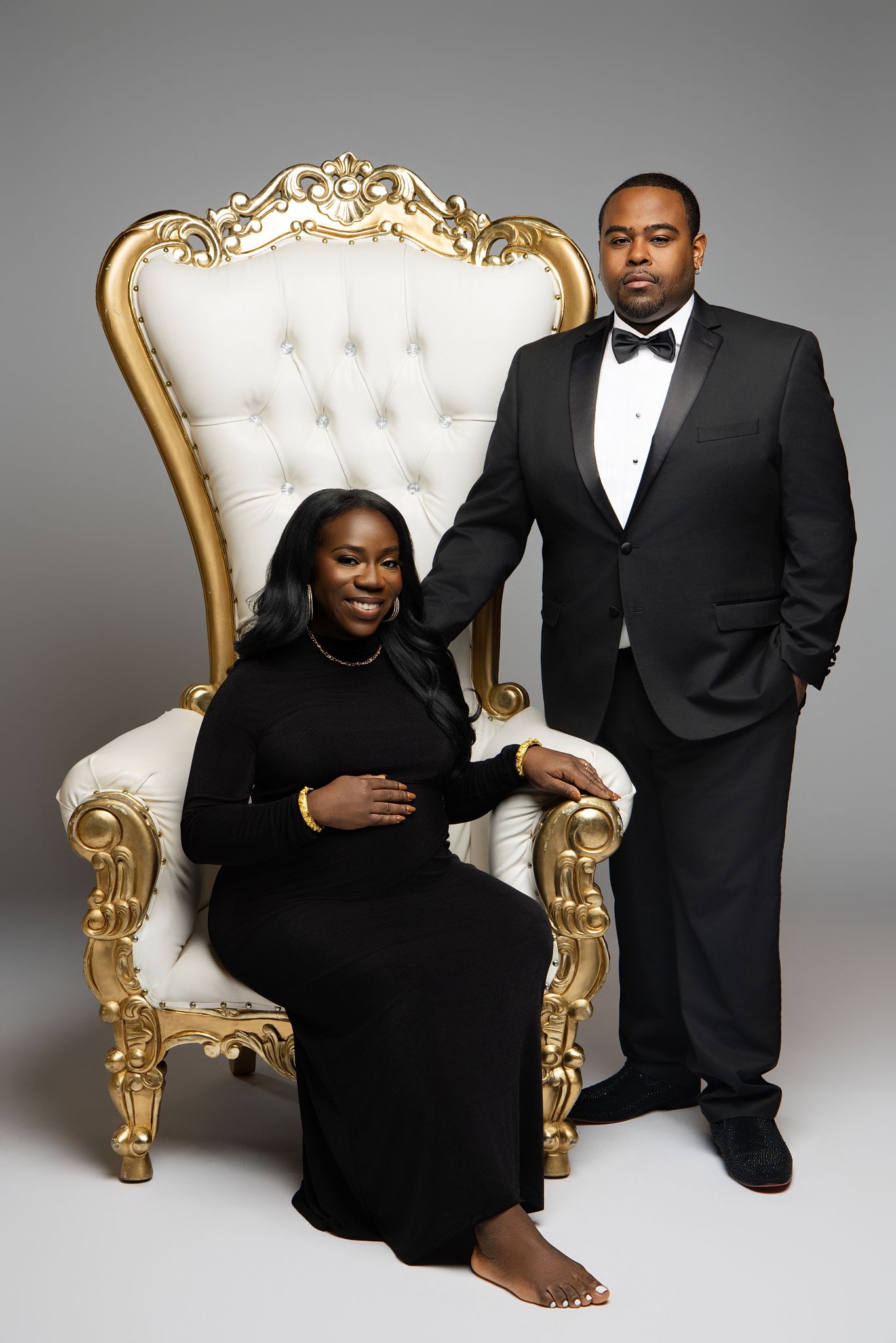 An elegant maternity portrait with a couple in black formal wear posed with an ornate gold and white throne chair.