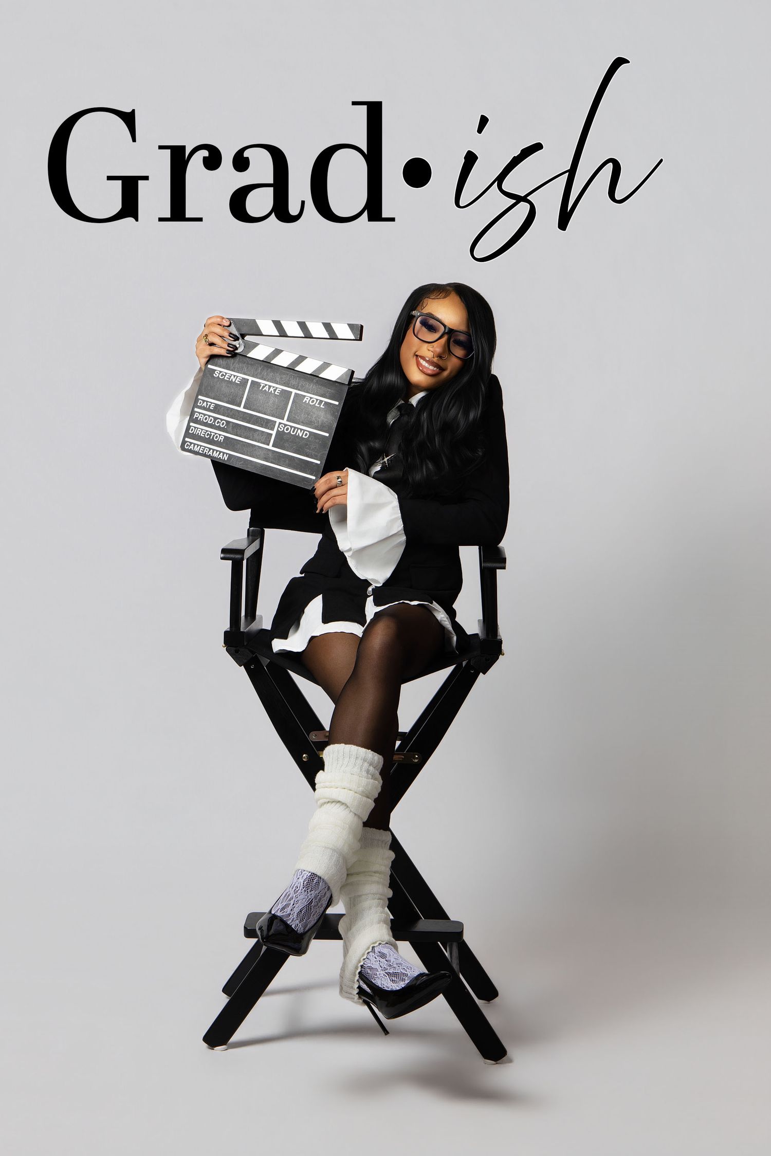 A graduate sits in a director's chair holding a clapperboard against a gray background in a stylish graduation photo.