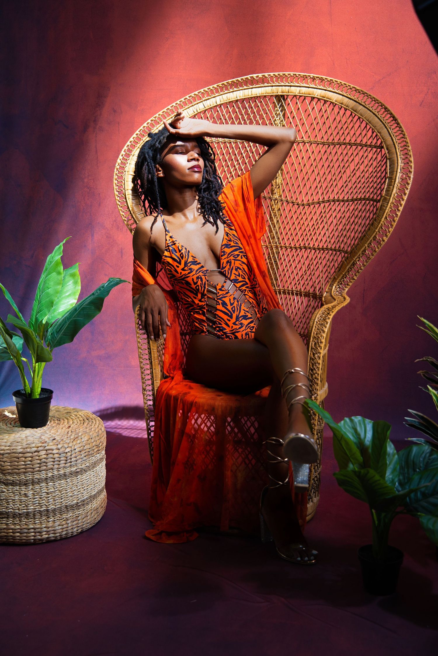 Model in orange dress poses on peacock chair with tropical plants against red backdrop.